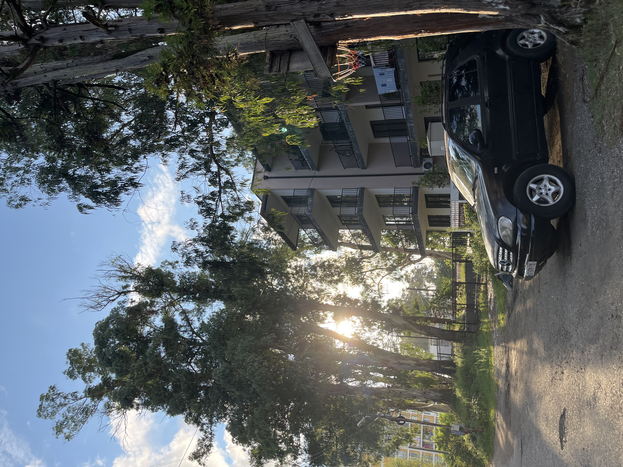 The image depicts a residential building and a parked car in Ureki, Georgia. A black SUV is parked on a dirt and asphalt road. The building is a multi-story structure with balconies and appears to be an apartment complex. Tall trees with sparse foliage surround the scene. The sun is shining, casting shadows and creating lens flares, indicating it is either morning or late afternoon. A basketball hoop is attached to a tree on the right side of the image. There are no people visible in the frame. The overall scene suggests a quiet, possibly suburban or semi-rural setting within Ureki.