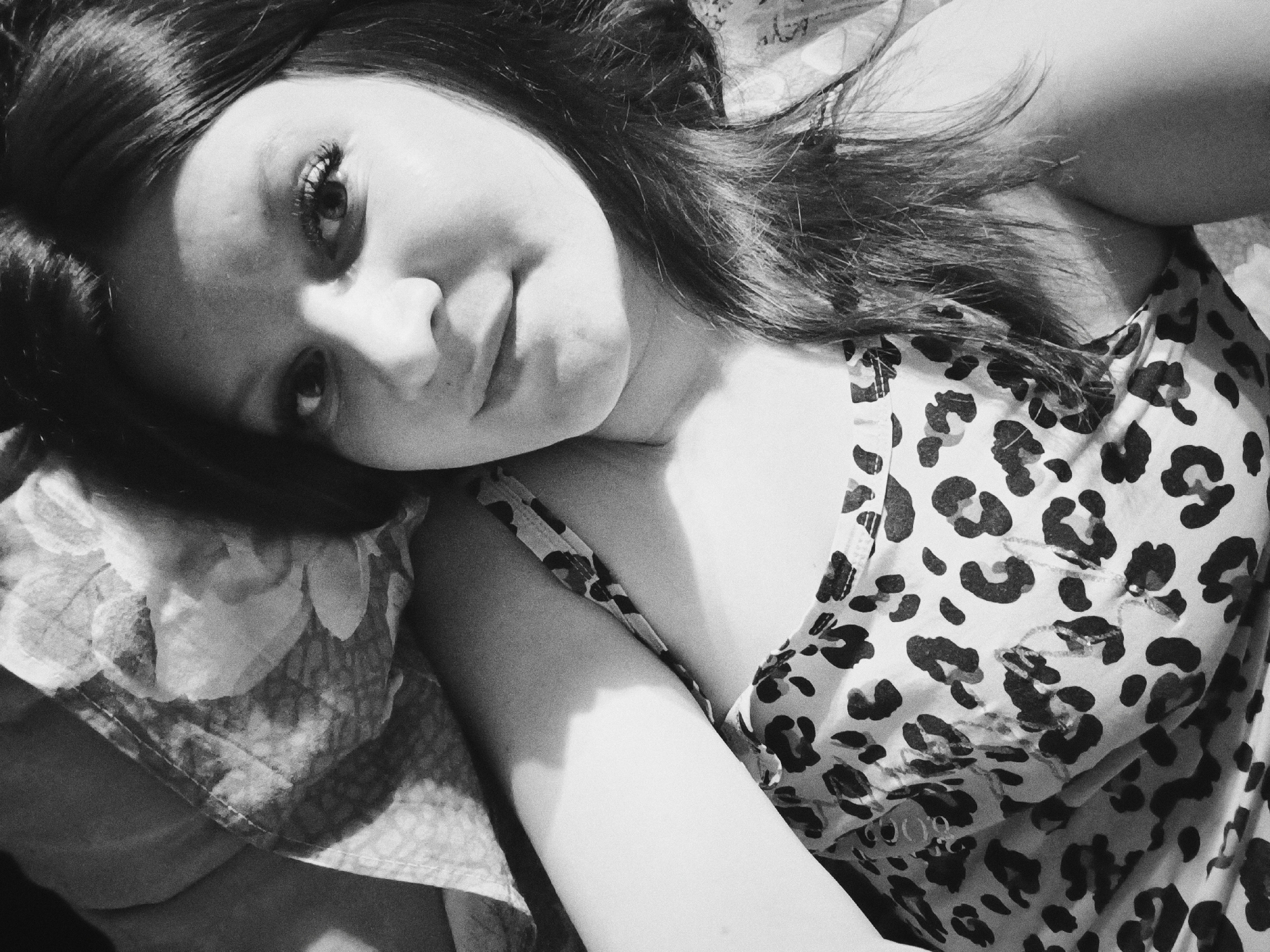 A black and white, close-up image shows a young woman reclining, her head resting on a floral-patterned pillow. She is wearing a leopard print top. Her gaze is directed toward the viewer. The lighting is uneven, creating strong shadows on her face and upper body. The location is not discernible from the image.
