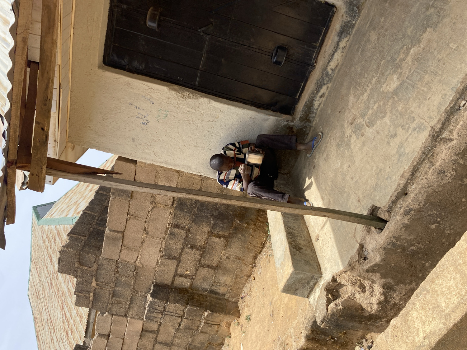 A man sits on the steps of a building, holding a container in his hands. The building has a dark door and a white wall with some inscriptions. The scene is in Barakin Akawo, Nigeria.