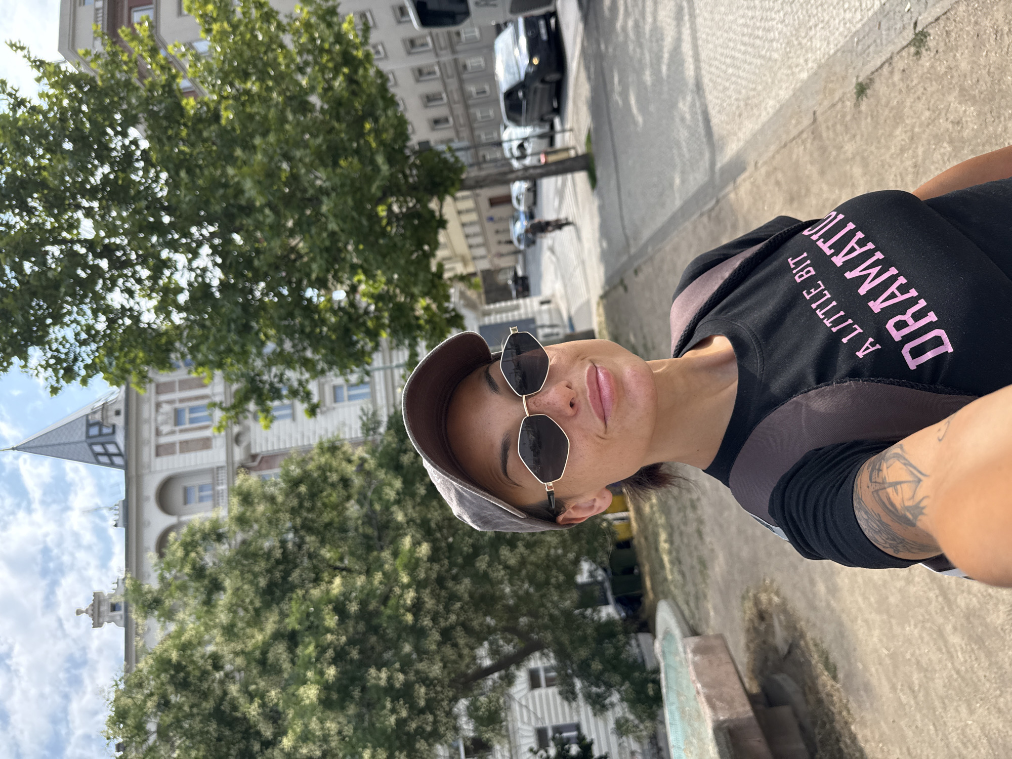 A woman is taking a selfie outdoors with a street and buildings in the background. She is wearing a dark t-shirt with pink lettering that reads "A LITTLE BIT DRAMATIC", a grey baseball cap, and sunglasses. She has a backpack on. In the background, there are trees, a building with a spire, and a street with cars and a person on a scooter. The scene is likely a public area in a city. Based on the architectural style of the building and the presence of a tram line (implied by the bus), this scene is consistent with an urban environment in Prague, Czechia, specifically near a building with a distinctive spire and ornate facade, possibly in the Vinohrady district.