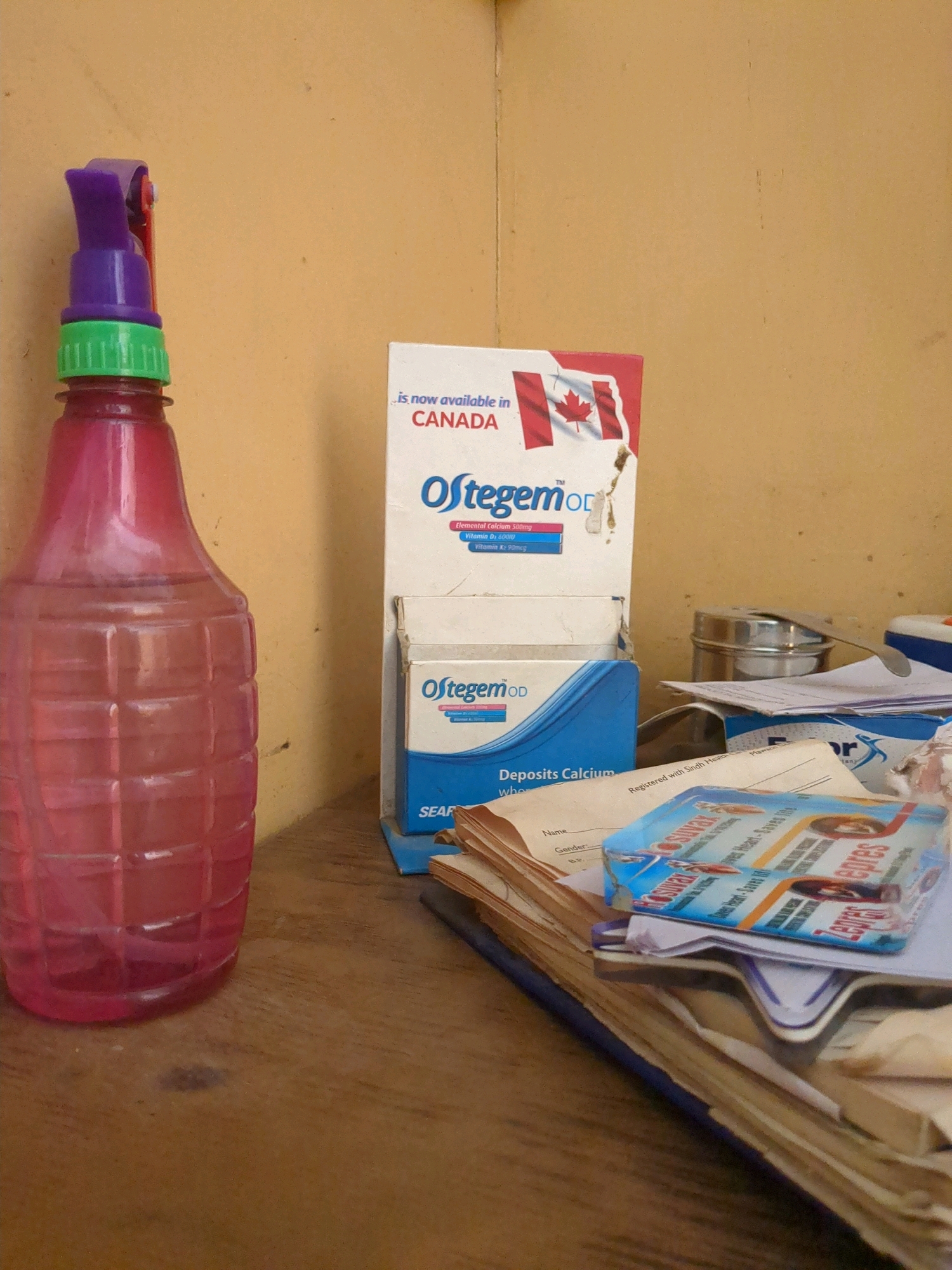 The image shows a close-up view of a wooden surface with various objects placed on it. In the foreground, on the left, is a pink plastic spray bottle with a purple and green trigger mechanism. To the right of the spray bottle, there are several pharmaceutical boxes stacked. One box, predominantly white with a red and white Canadian flag motif, reads "Ostegem OD" and mentions it is "now available in CANADA." Below this, another blue and white box also says "Ostegem OD" and has the text "Deposits Calcium." Further to the right and in the background, there is a silver tin can and some papers. A stack of papers with some writing, including "SEAR," is visible in front of the blue and white box. On top of the stack of papers, a brightly colored packet, possibly of medication or a health product, is placed. The lighting suggests an indoor environment with a warm yellow wall in the background. The overall scene appears to be a collection of items on a desk or shelf. There is no discernible information within the image to indicate the location within the city of Karachi Division, Pakistan.