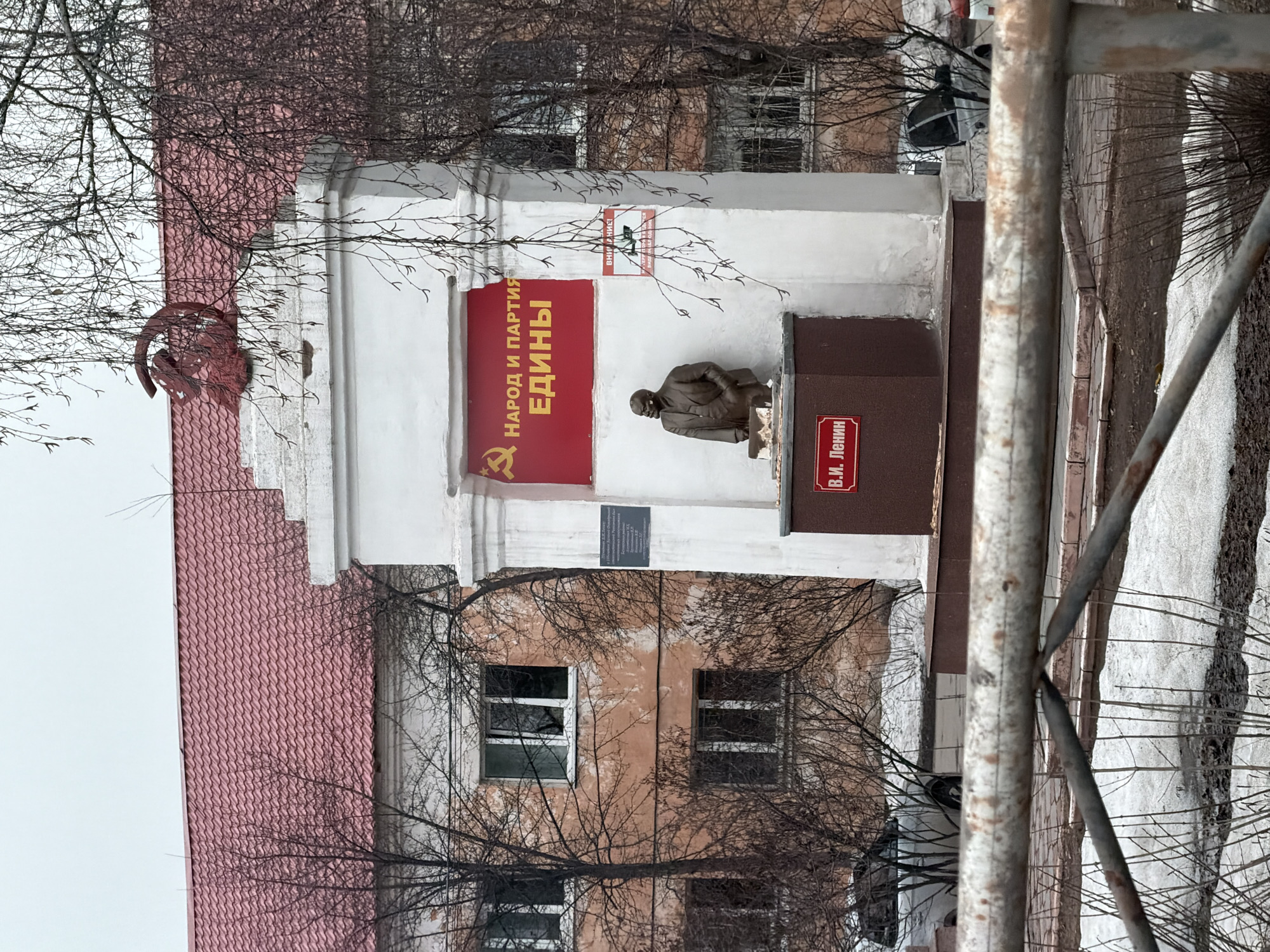 The image depicts a monument to V.I. Lenin located in Beloyarsky, Russia, observed under an overcast sky. The central feature is a white, multi-tiered concrete monument topped with a prominent red hammer and sickle emblem. Directly below the emblem, a red banner displays the yellow Russian text "НАРОД И ПАРТИЯ ЕДИНЫ" (The People and Party are United), accompanied by a smaller hammer and sickle symbol. A bronze bust of V.I. Lenin, portrayed from the waist up in a suit with his left hand extended, is mounted on a dark brown pedestal at the base of the white structure. A red plaque on this pedestal reads "В.И. Ленин". A rectangular dark plaque is affixed to the left side of the white monument, and a smaller sign with green elements is positioned on its right side. In the background, a multi-story building with a faded peach-colored facade, multiple white-framed windows, and a red corrugated metal roof is visible. Bare deciduous trees with sparse branches partially obscure the background building and frame the monument. The ground exhibits patches of melting snow interspersed with damp, dark earth. A light-colored vehicle is partially visible parked behind the monument on the left. A rusty metal barrier is present in the immediate foreground, horizontally traversing the lower portion of the frame.