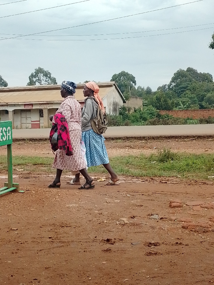 Two women, one wearing a pink and white dress and a blue head covering, the other wearing a blue and white dress and an orange head covering, walk along a dirt road in Kivaywa, Kenya. The road is muddy, and there is a green sign in the foreground. The location is near the junction of the Kivaywa-Nambale road and the Kivaywa-Kisumu road. Behind the women, there are buildings with a red-tile roof, trees, and a brick wall. There are power lines overhead.