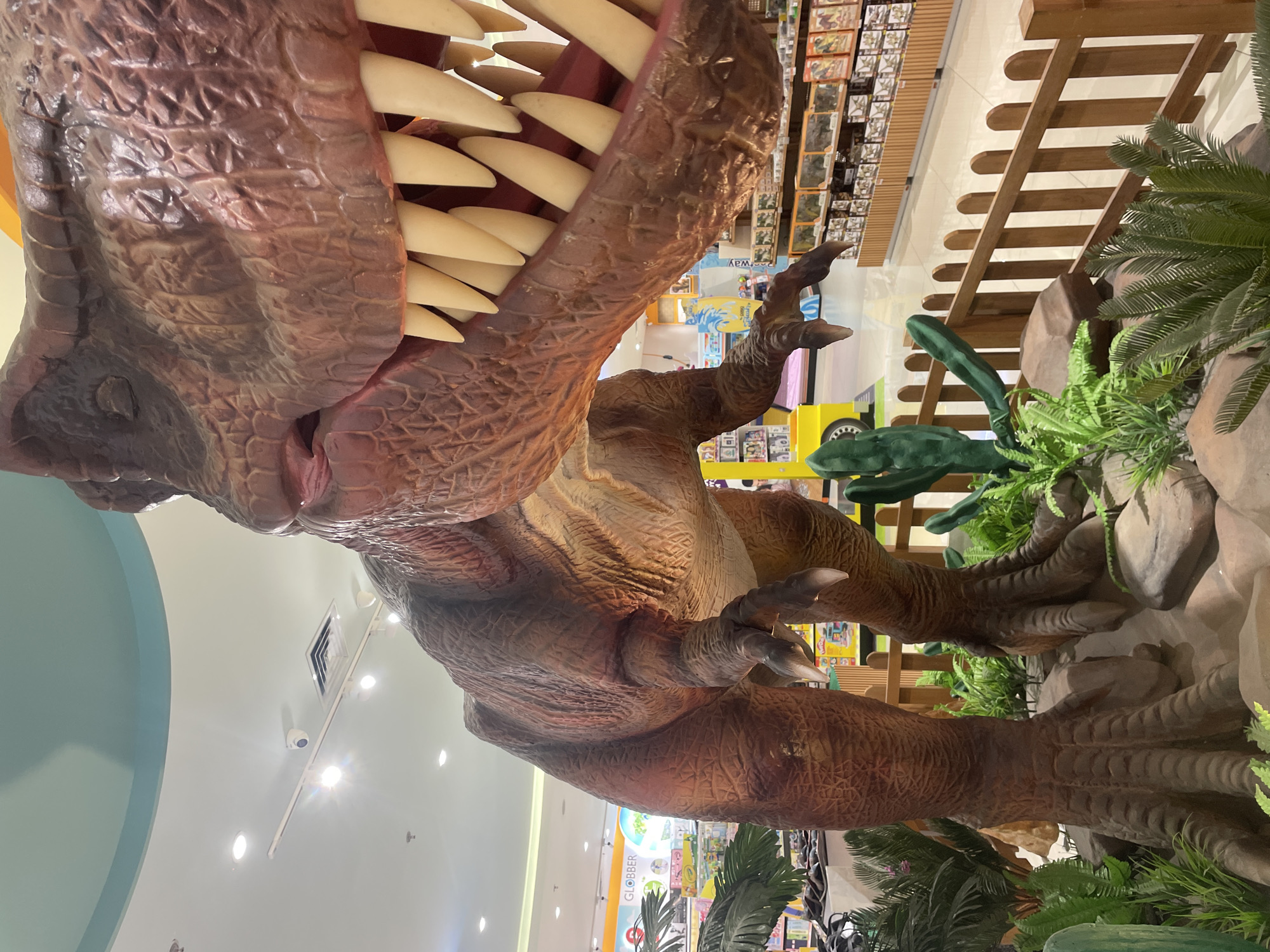 The image shows a large, animatronic Tyrannosaurus Rex display inside a building, possibly a retail or entertainment venue. The T-Rex is brown, with many exposed, pointed, white teeth. The dinosaur is surrounded by artificial plants and faux rocks. A wooden fence and shelves stocked with merchandise are visible in the background. Based on the presence of "Globber" branding, this image was likely taken at Level 21 Mall Denpasar, Indonesia.