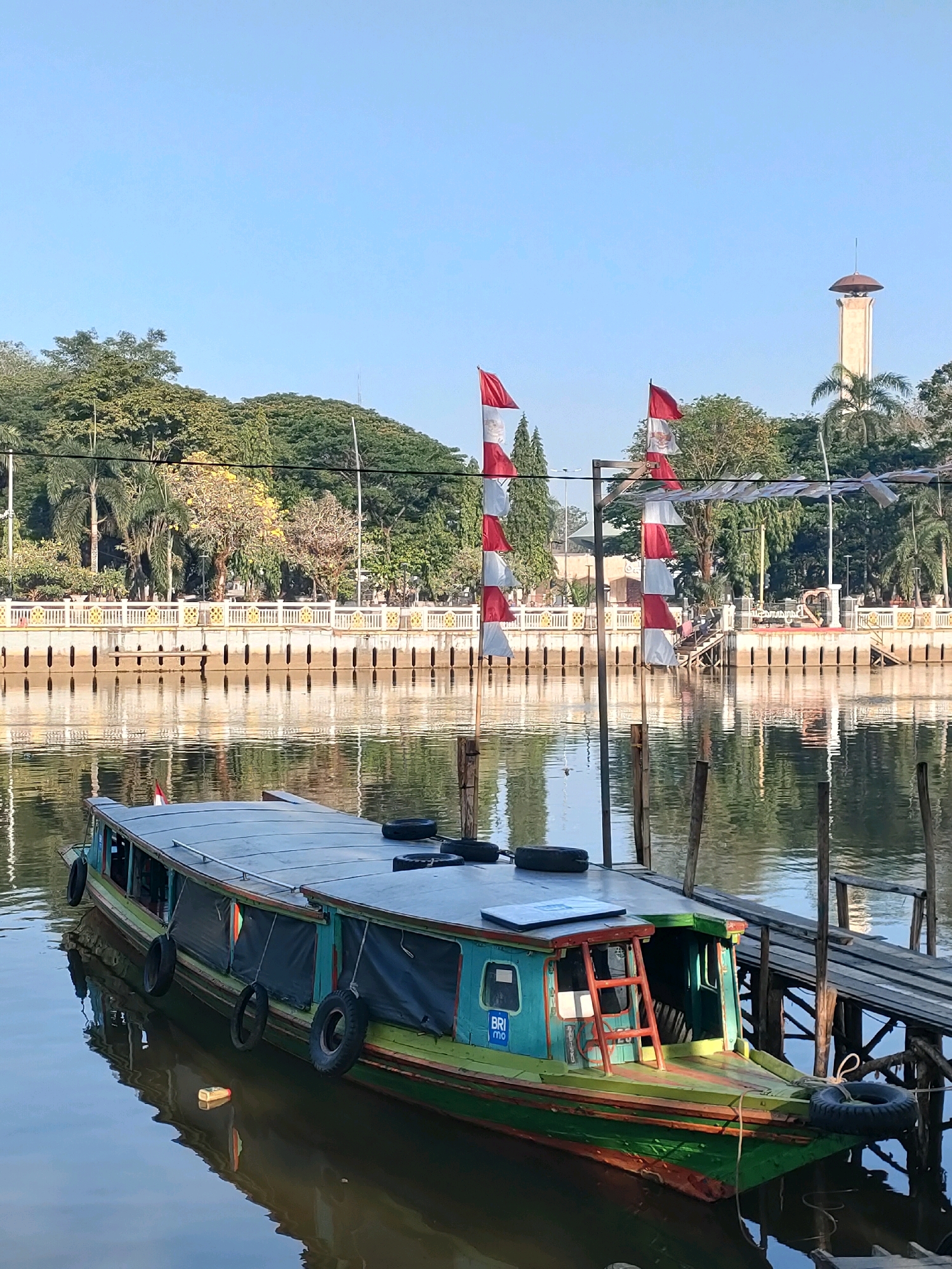 The image captures a serene scene of a river in Banjarmasin, Indonesia, likely during the morning given the clear blue sky and soft lighting. In the foreground, a long, narrow boat with a covered roof is docked next to a wooden pier. The boat is painted in vibrant colors, primarily green and blue, with red accents. Several black tires are affixed to its sides, serving as fenders. The water reflects the boat, the pier, and the surrounding environment, creating a mirrored effect.
Beyond the river, a well-maintained embankment is visible, lined with a decorative white fence featuring intricate gold patterns. Behind the fence, lush green trees and some yellow flowering trees create a verdant backdrop. In the distance, a tall, slender tower with a domed top stands against the sky, possibly a minaret or a landmark. Two flagpoles with alternating red and white Indonesian flags are prominent, adding a patriotic touch to the scene. The overall impression is one of a peaceful urban waterfront, showcasing a blend of natural beauty and human infrastructure.