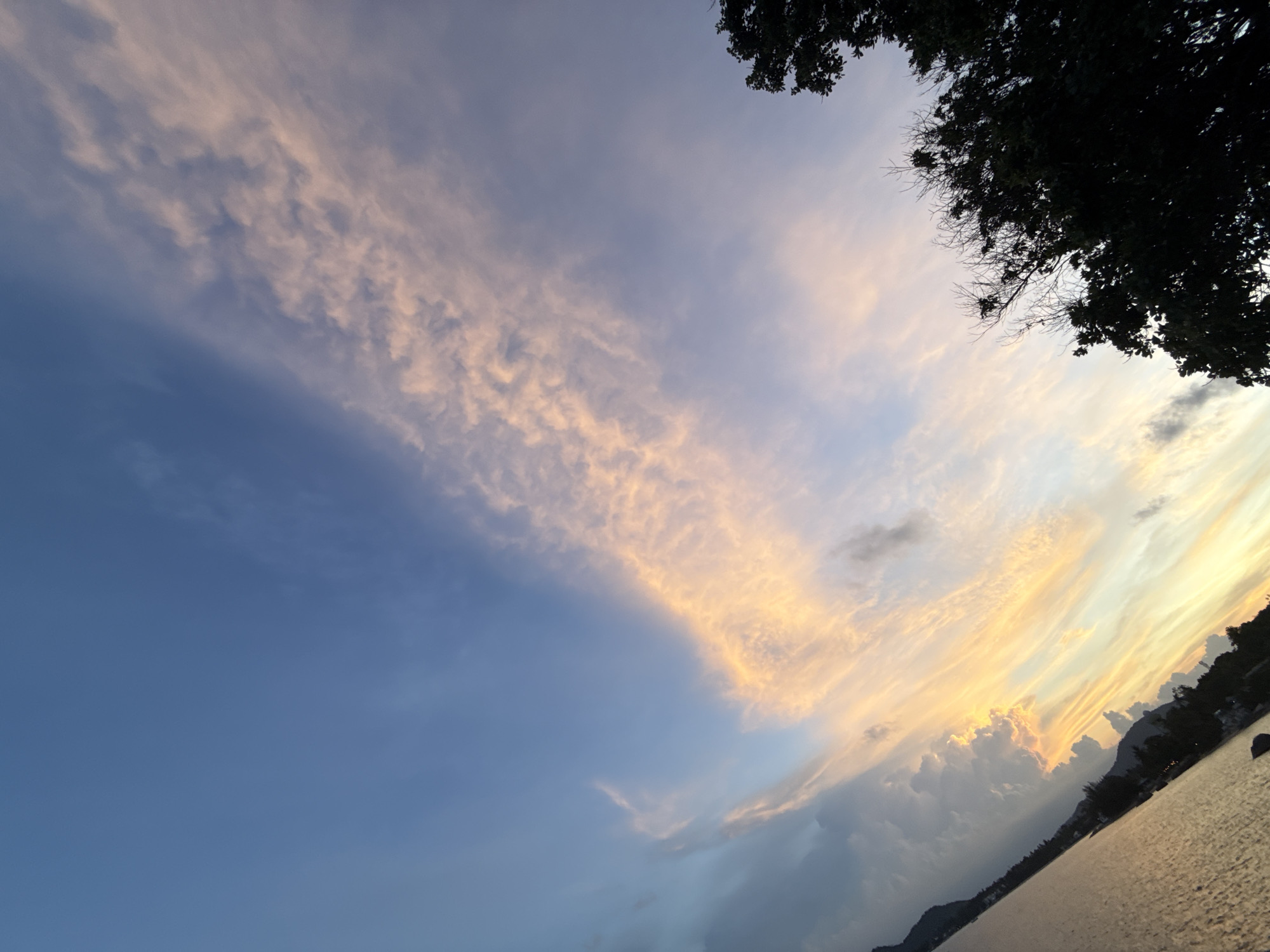 The image depicts a scene in Baan Hua Thanon, Thailand, during what appears to be dusk or dawn. The sky is a gradient of blue, transitioning into hues of soft orange and pink, illuminated by the setting or rising sun. Wispy clouds are scattered across the sky, some catching the light and appearing in shades of peach and gold. In the lower left portion of the image, a body of water, likely the sea, reflects the warm colors of the sky. Along the horizon, silhouetted landforms, possibly hills or mountains, are visible, as are dark shapes of trees and vegetation. The overall impression is one of natural beauty and tranquility. There are no people or specific objects of interest readily identifiable in the foreground or midground.
