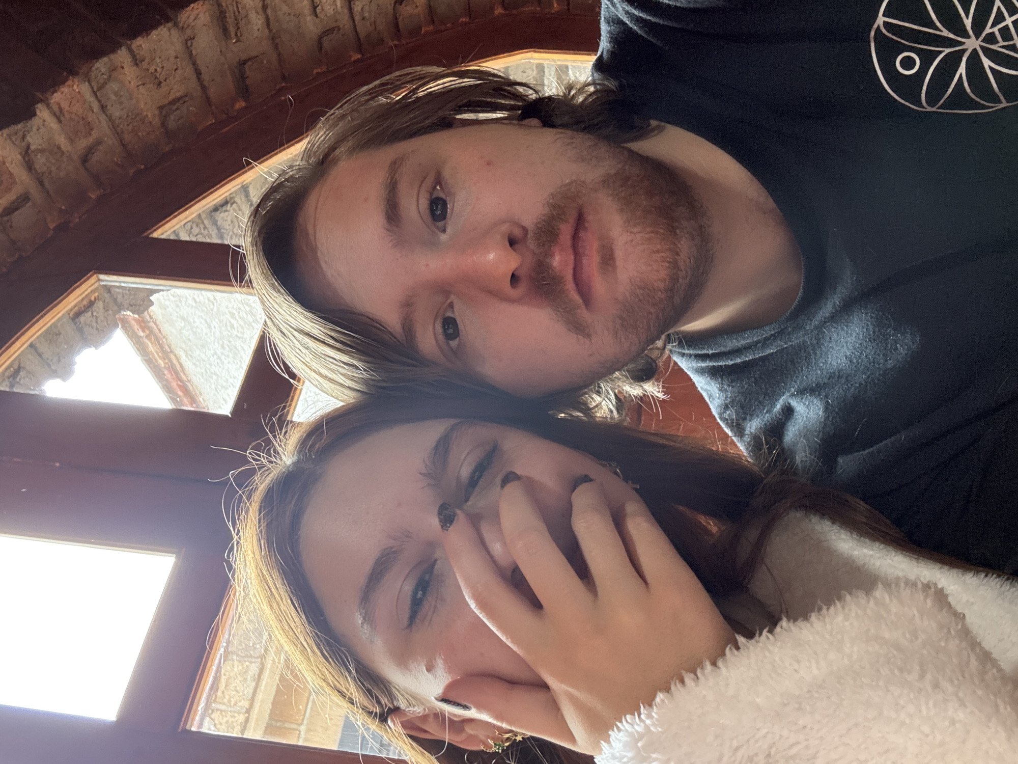 The image is a selfie of a man and a woman, possibly taken in Cañada de Madero, Mexico, though no specific landmarks are visible. The man is positioned slightly above and behind the woman. He has light brown hair and a beard. He is wearing a dark t-shirt with a circular design on the upper right. The woman is in the foreground, with light brown hair and is wearing a white, fuzzy jacket. She has one hand partially covering her face. The background consists of an interior space with a window showing a bright outdoor scene and the ceiling is decorated with what appears to be brick.