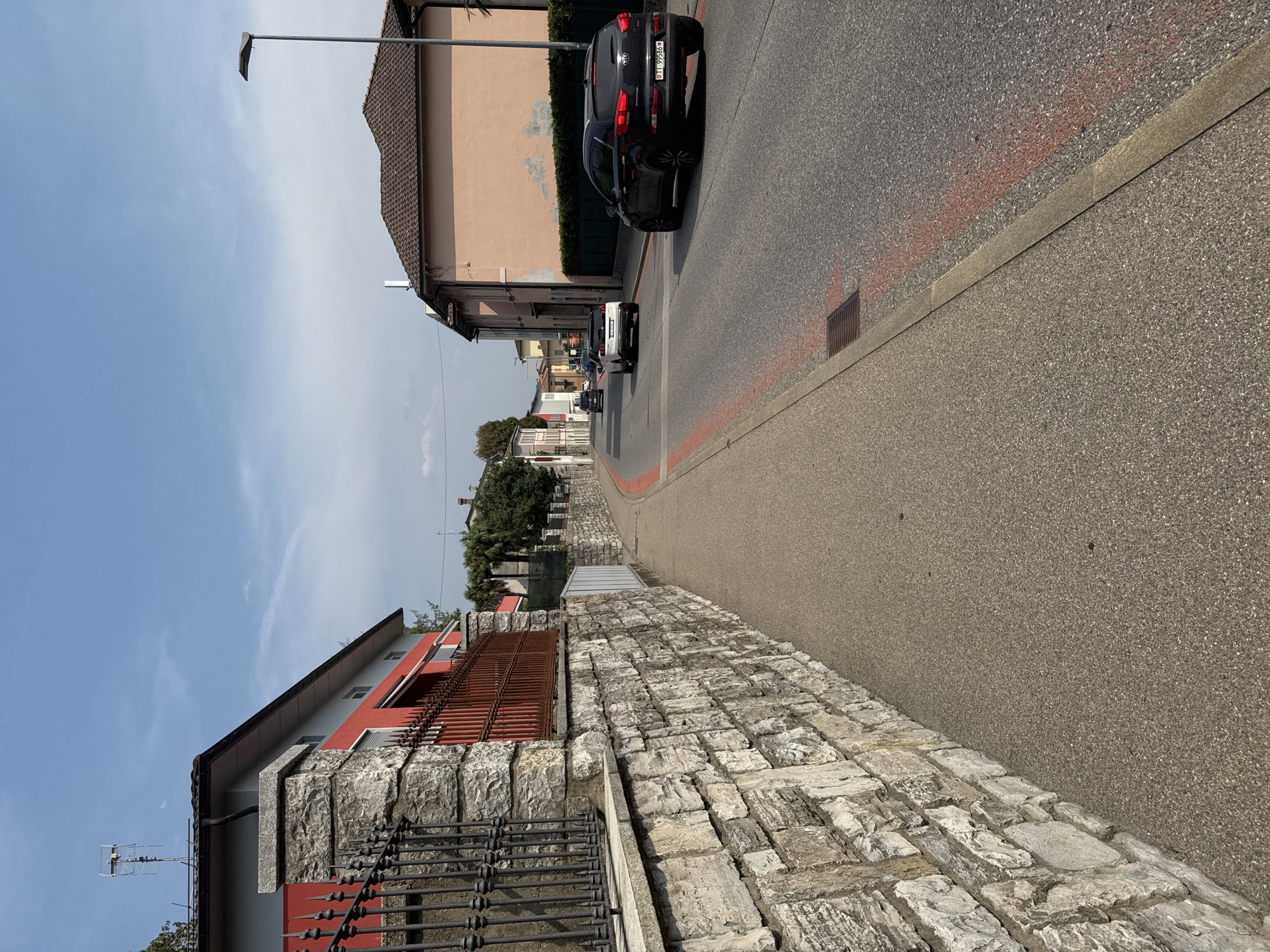 The image depicts a street scene in Balerna, Switzerland. On the left, a low stone wall borders a grey asphalt sidewalk. The wall is topped with a black metal fence and a red-painted railing. Behind the wall is a building with a red facade and grey upper sections, topped with a grey roof and a television antenna. On the right side of the street, a peach-colored building with a tiled roof stands adjacent to the road. A streetlamp is positioned in front of this building. The road itself is asphalt with red painted markings. Several vehicles are visible: a black Kia Sportage with a Swiss license plate (TI 99586) is in the foreground on the right, a white Peugeot is driving away in the middle distance, and another black car is further down the road. Palm trees and other greenery are visible along the right side of the street. The sky is blue with scattered white clouds. The scene appears to be set during the daytime.