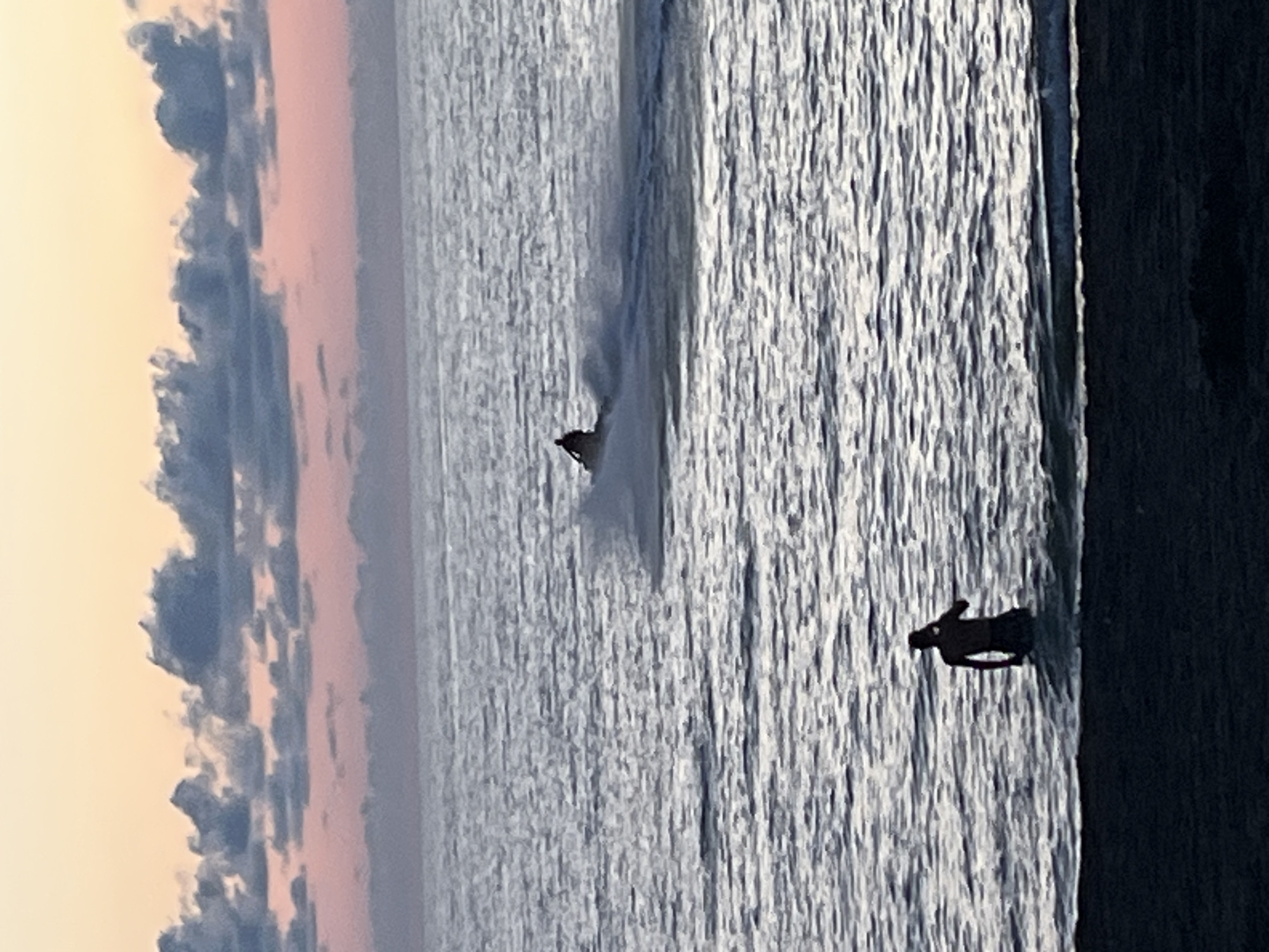 The image shows a seaside scene at dusk. In the foreground, a dark, textured area, likely sand or beach, borders the water. A person, silhouetted and appearing to be a male, stands partially submerged in the water, facing away from the camera and towards the sea. Further out in the water, a jet ski is being ridden, creating a spray of water as it moves across the surface. The sky above is a gradient of soft colors, transitioning from a peachy-orange near the horizon to a pinkish-purple and then a pale blue, with scattered, dark clouds. The sea itself is rendered in muted tones of gray and blue, with ripples and light reflections indicating movement and shallow depth. The overall impression is one of a peaceful evening by the Black Sea coast in Ureki, Georgia.