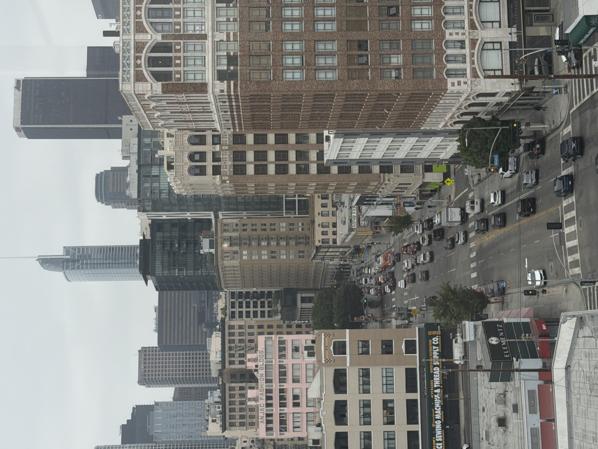 This high-angle, slightly rotated image presents a dense urban landscape, likely an aerial view looking down onto a city street. The scene is dominated by numerous multi-story buildings, showcasing a mix of architectural styles, from modern glass high-rises to older, intricately designed brick and stone structures.
The street below is a multi-lane thoroughfare, bustling with vehicular traffic. Many cars and trucks are visible, either moving or stopped in lanes, and pedestrian crosswalks and street markings are evident. Traffic lights are also visible at intersections. Scattered trees punctuate the concrete environment, adding touches of greenery.
The time of day appears to be daytime, under an overcast or cloudy sky, as there are no strong shadows. The overall impression is one of a busy commercial and residential district.
Visible text on buildings and signs includes: "ANJAC FASHION BLDGS." on a pink building, "LOFTS," "ELEMENTZ," and "PEGASUS WHOLESALE DISTRIBUTOR." A sign for "CE SEWING MACHINE & THREAD SUPPLY CO.," along with "RESSFOR IRONS" and "SUNDORS & CUTTIN STEAMERS," is also present. A street sign reads "Maple," and Korean text "미상백화" is also visible. This urban setting is consistent with a major city like Los Angeles, United States of America.