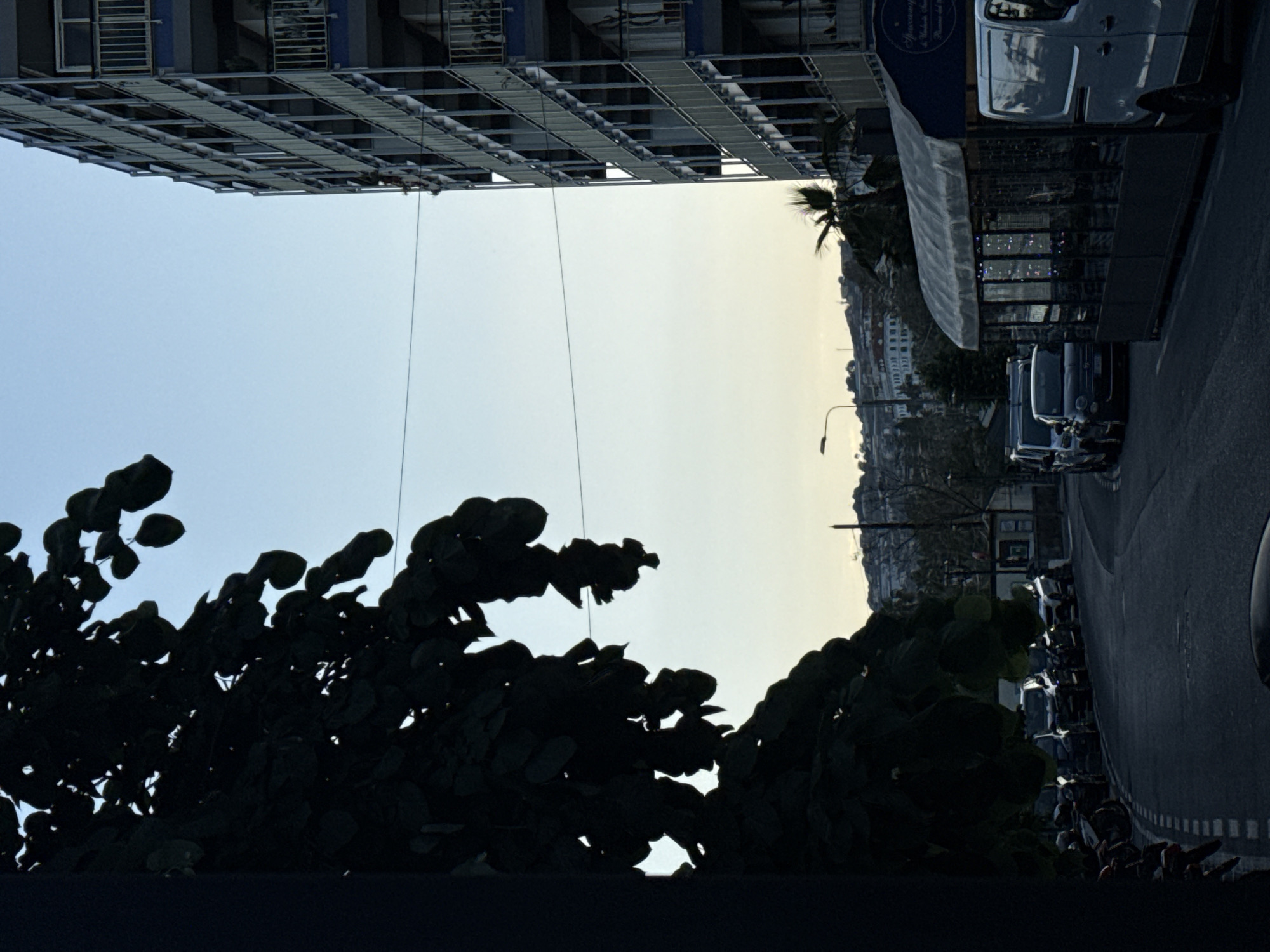 This is an outdoor scene in Sanremo, Italy, with a high-rise building under construction on the left. The sky transitions from blue to a pale yellow-white, indicating either sunrise or sunset. Dark foliage dominates the lower portion of the image, silhouetted against the sky. On the right side of the image, the view shifts to a street with parked vehicles and a storefront with a blue awning. Further into the distance, there are buildings on a hillside. A street lamp is visible near the street.