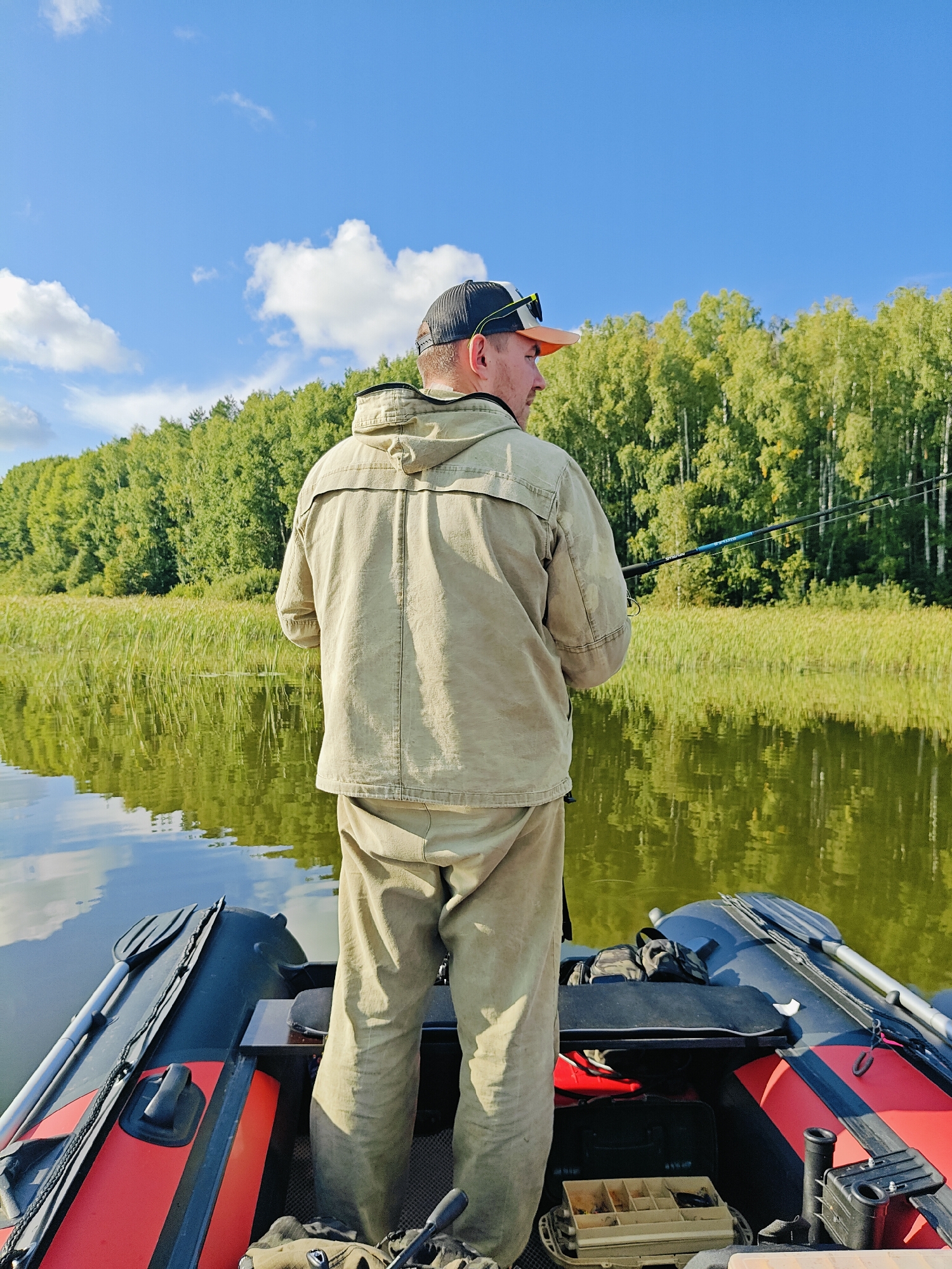 A man stands in an inflatable boat, fishing on a body of water. He is wearing a light-colored jacket and pants, and a baseball cap. He is holding two fishing rods. The boat is equipped with oars and fishing gear, including a tackle box. The background features a dense forest with green trees and a blue sky with white clouds. The water is calm and reflects the surrounding trees and sky. The scene appears to be in Chaykovsky, Russia, given the context of the query.