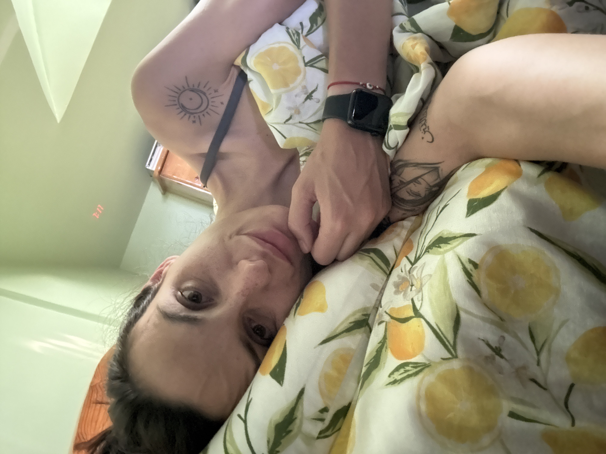 A selfie-style, medium-angle close-up depicts a person lying in bed covered with a lemon-patterned comforter. The individual has light skin, dark hair pulled back, and visible freckles. A black Apple Watch is on their left wrist, adorned with a red beaded bracelet. Tattoos are visible on the arms, including a sun and moon design, another on the upper arm, and one near the left elbow. The background suggests an indoor space with light green walls and a glimpse of a wooden cabinet. The picture appears to have been taken indoors, but information about the city of Pleven, Bulgaria, is not visible within the image.