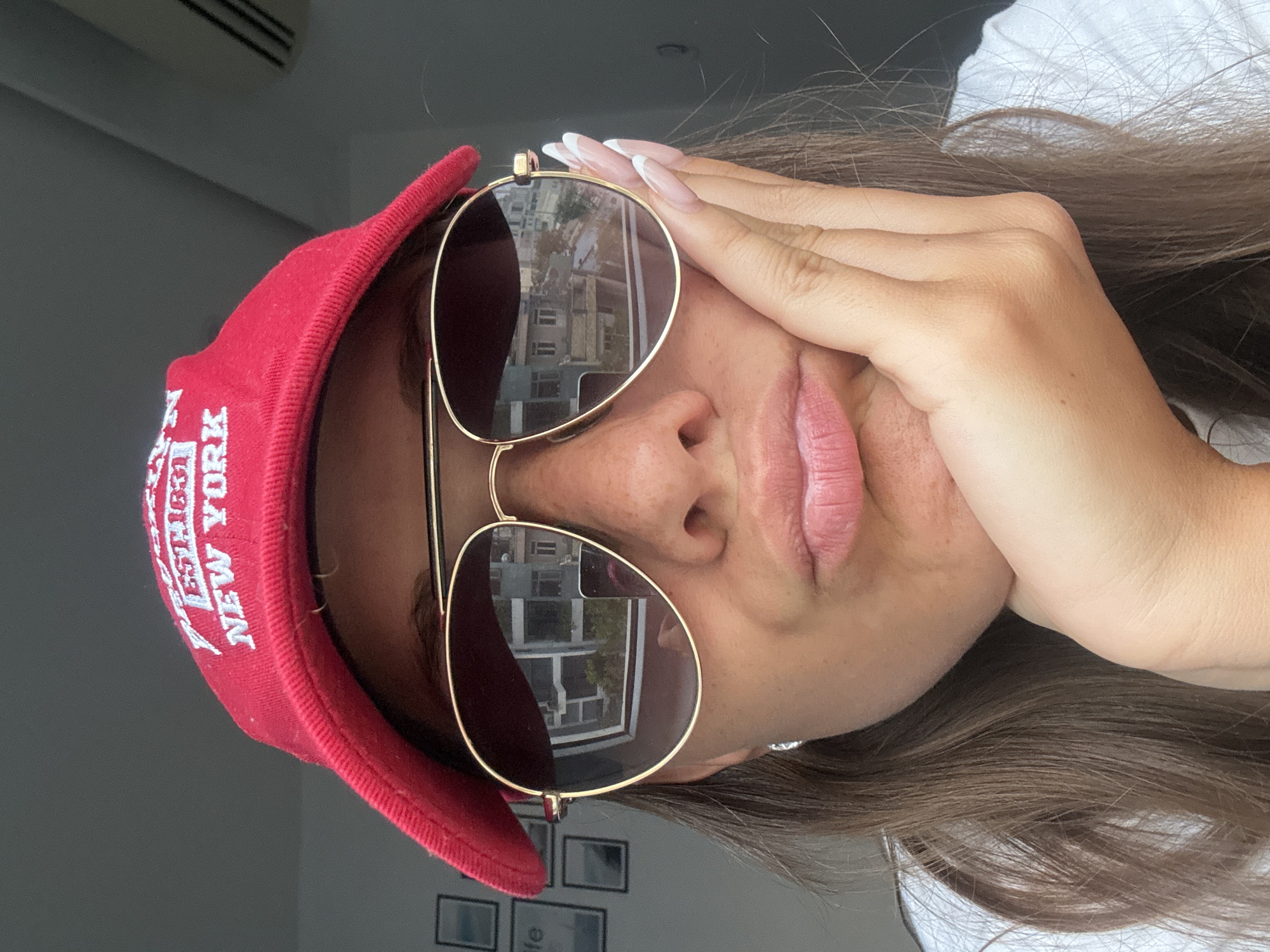 A woman is shown in a close-up, rotated so that the top of her head is facing right. She is wearing a red cap with the words "New York" embroidered on it, and gold-rimmed sunglasses that reflect a building and trees. Her right hand is partially covering her forehead. Her hair is brown and loose. The background includes a white wall and framed objects below. The location cannot be determined from the image.