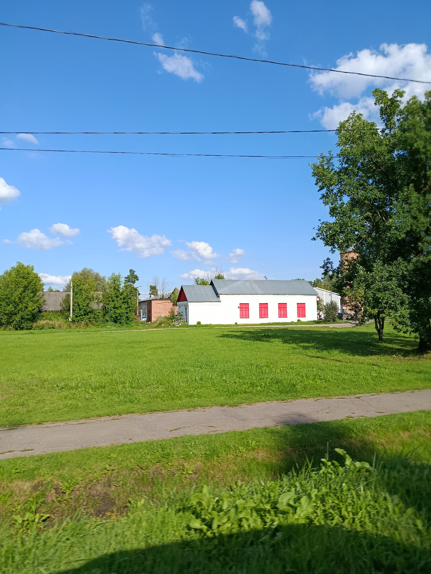 The image captures a sunny day in the city of Vereya, Russia, featuring a white building with a red-tiled roof. This structure has three red-framed windows visible on its side. In front of the building, a vibrant green lawn stretches out, and a paved pathway runs through it, casting shadows from unseen objects. To the right, a large, leafy green tree dominates the foreground, its branches reaching towards the clear blue sky dotted with fluffy white clouds. Beyond the white building, several other structures, including a brick building, are partially visible, interspersed with more greenery and trees. The overall scene depicts a peaceful, rural setting under a bright sky.