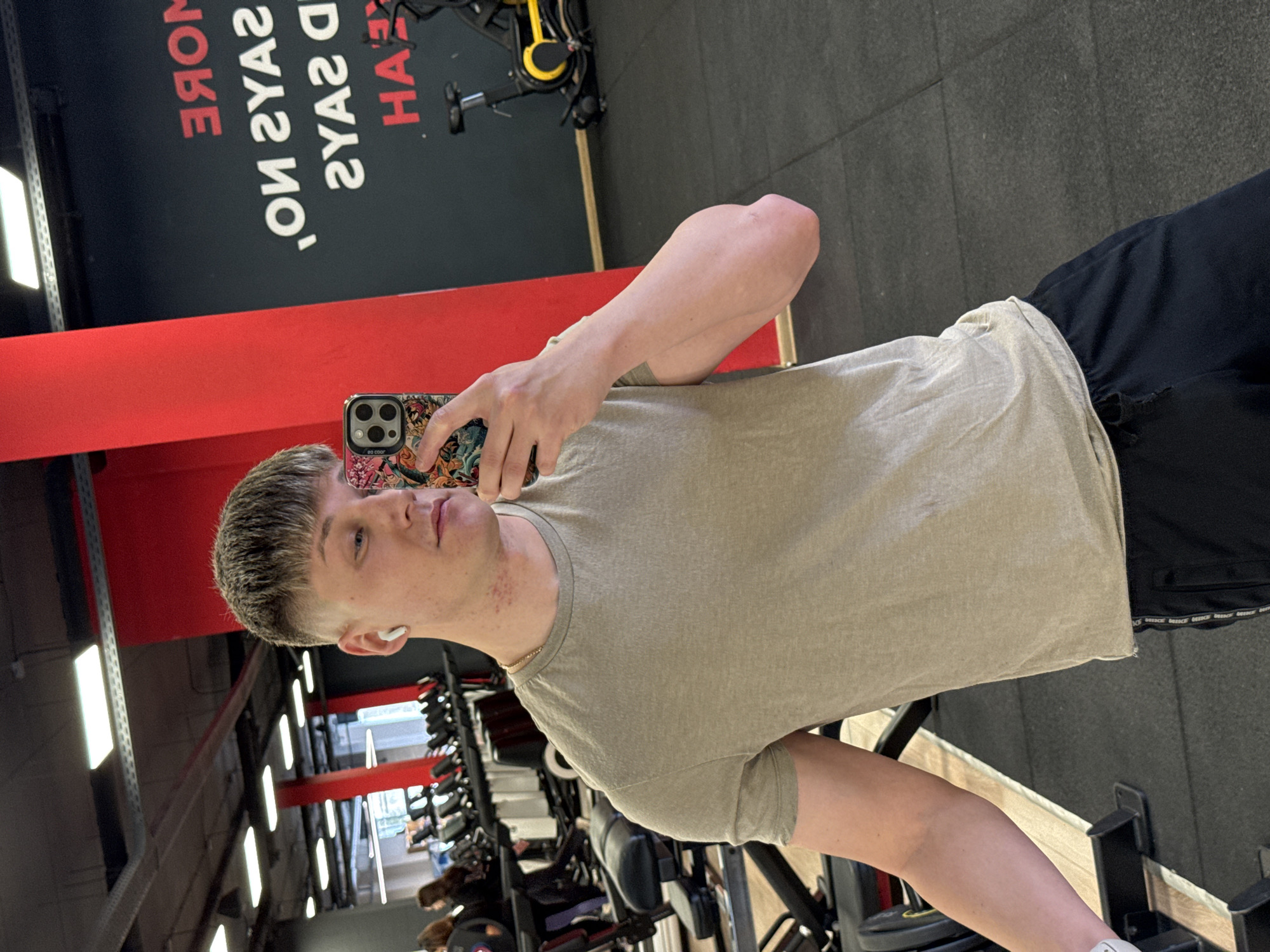 The image shows a young man indoors, likely in a gym in Plovdiv, Bulgaria. He is fair-skinned with short, light brown hair, wearing a beige t-shirt and black shorts. An earbud is visible in his left ear. He is holding a smartphone with a colorful case in his right hand, presumably taking a selfie. Gym equipment is visible in the background, including weight racks and exercise bikes. Motivational signage is visible on the wall behind him. The floor is dark, likely made of rubber or similar gym flooring material.