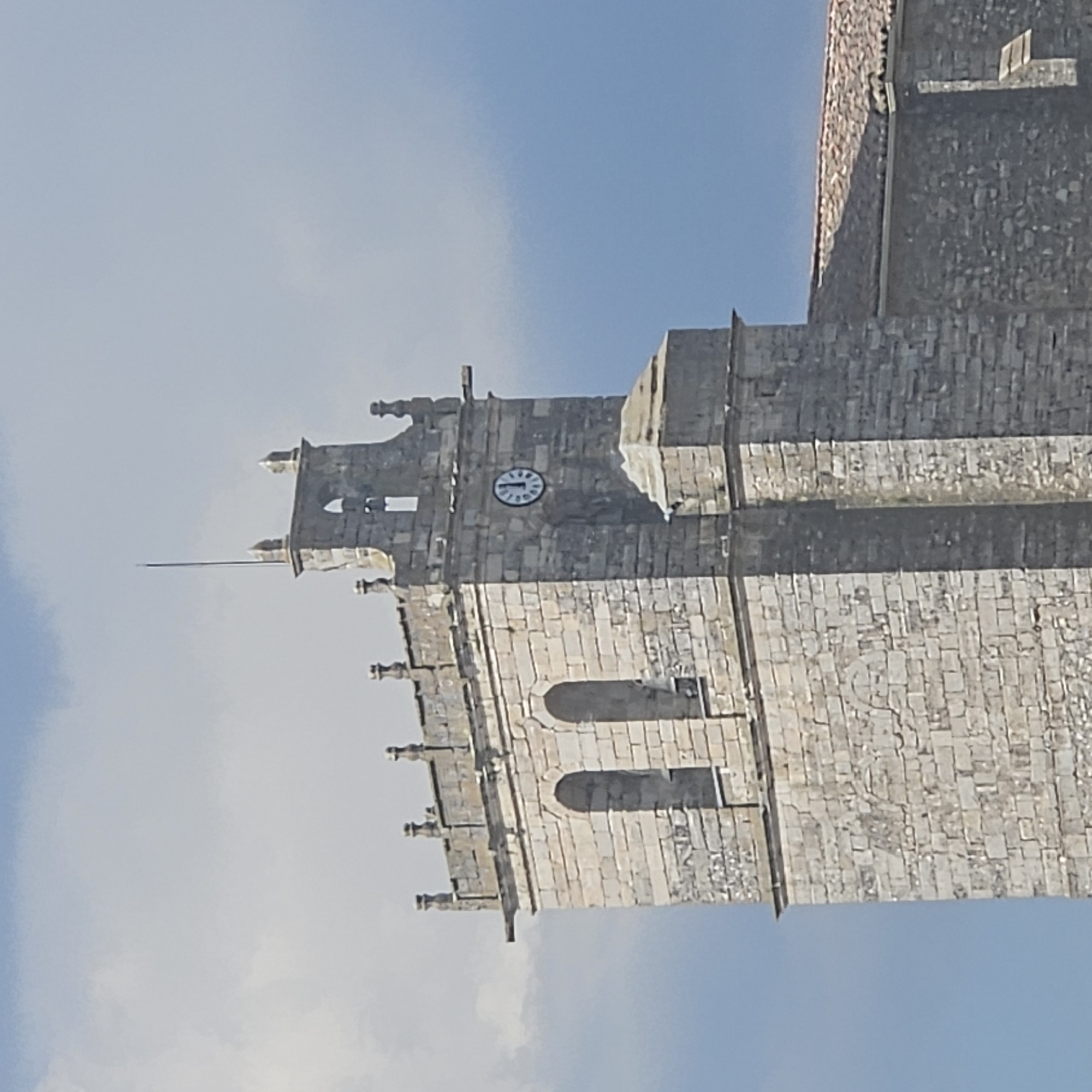 A stone tower with a clock on one side is visible against a bright blue sky. The clock shows approximately 11:57. The tower is constructed of light-colored stone blocks and features several architectural details including windows and gargoyles. A needle or spike extends from the top of the tower. The image appears to be taken at an angle, with the tower extending diagonally from the lower right to the upper left. The location is Algora, Spain.