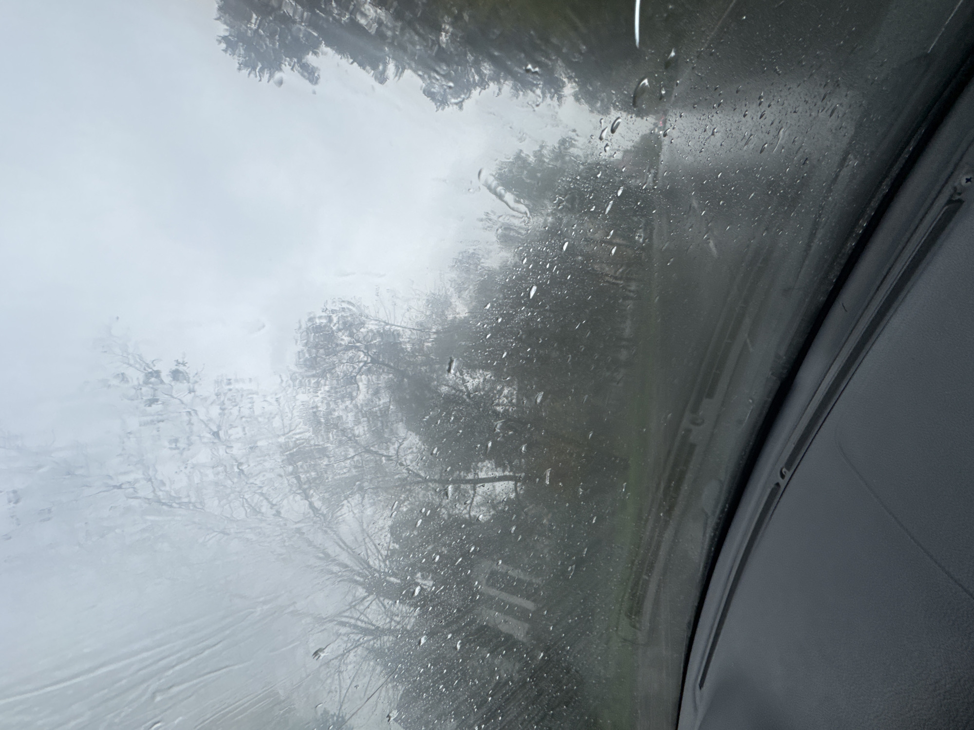 The image captures a view through a car windshield on a rainy day. The windshield is covered in raindrops, distorting the view of the outside world. In the background, trees with bare branches are visible, suggesting it is likely autumn or winter. The overall atmosphere is grey and muted, typical of overcast weather. The scene appears to be a road with other vehicles in the distance. The photograph is taken from inside a car, looking out at the street. There are no people clearly visible in the image, and the focus is on the weather conditions and the environment. It is not possible to determine the specific location in the city of Perm, Russia from the provided image.