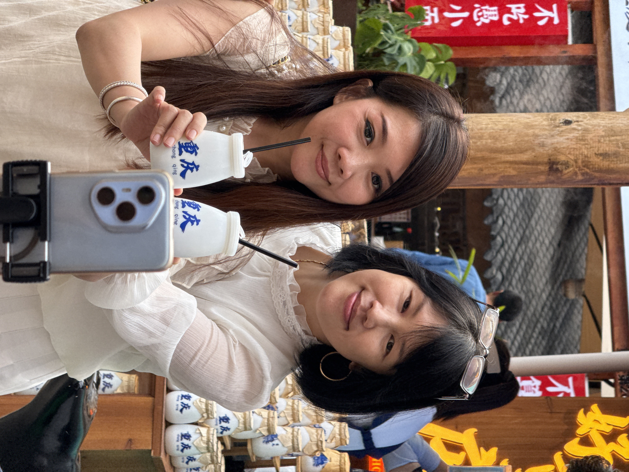 Two smiling Asian women are taking a selfie with a gray smartphone. The woman on the left has black hair and is wearing a white lace-trimmed blouse and large gold hoop earrings; she is holding a white bottle with a blue label that reads "重庆" (Chongqing). A black straw is visible in her bottle. The woman on the right has long brown hair and is wearing a light tan dress; she is also holding a bottle of the same type. They are surrounded by shelves stacked with more of the labeled bottles. The background includes red signs with white Chinese characters and some greenery. The scene appears to be outdoors or near a window, with wooden architectural elements visible. The bottles are from Chongqing. Based on the style of architecture and the writing on the signs, the location is likely within the Yuzhong District in China.