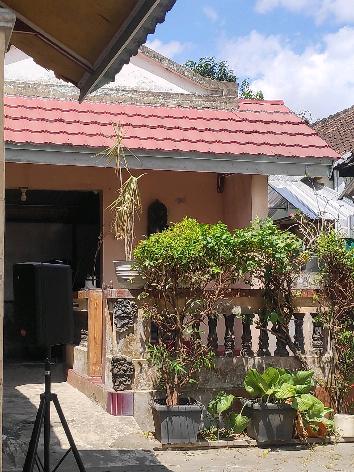 The image shows a close-up view of the exterior of a house on a sunny day. The house has a red tiled roof and is painted in earthy tones. In the foreground, there is a black speaker on a tripod, positioned on a concrete patio. To the right of the speaker, there is a decorative stone railing with carved lion-head motifs. Several potted plants are arranged along the railing, including a lush green plant in a dark pot in the center. Behind the railing, a portion of the house's facade is visible, with a window or open doorway. The scene appears to be set in a residential area in Kebonarum, Indonesia, given the architectural style and lush vegetation. The sky above is partly cloudy.