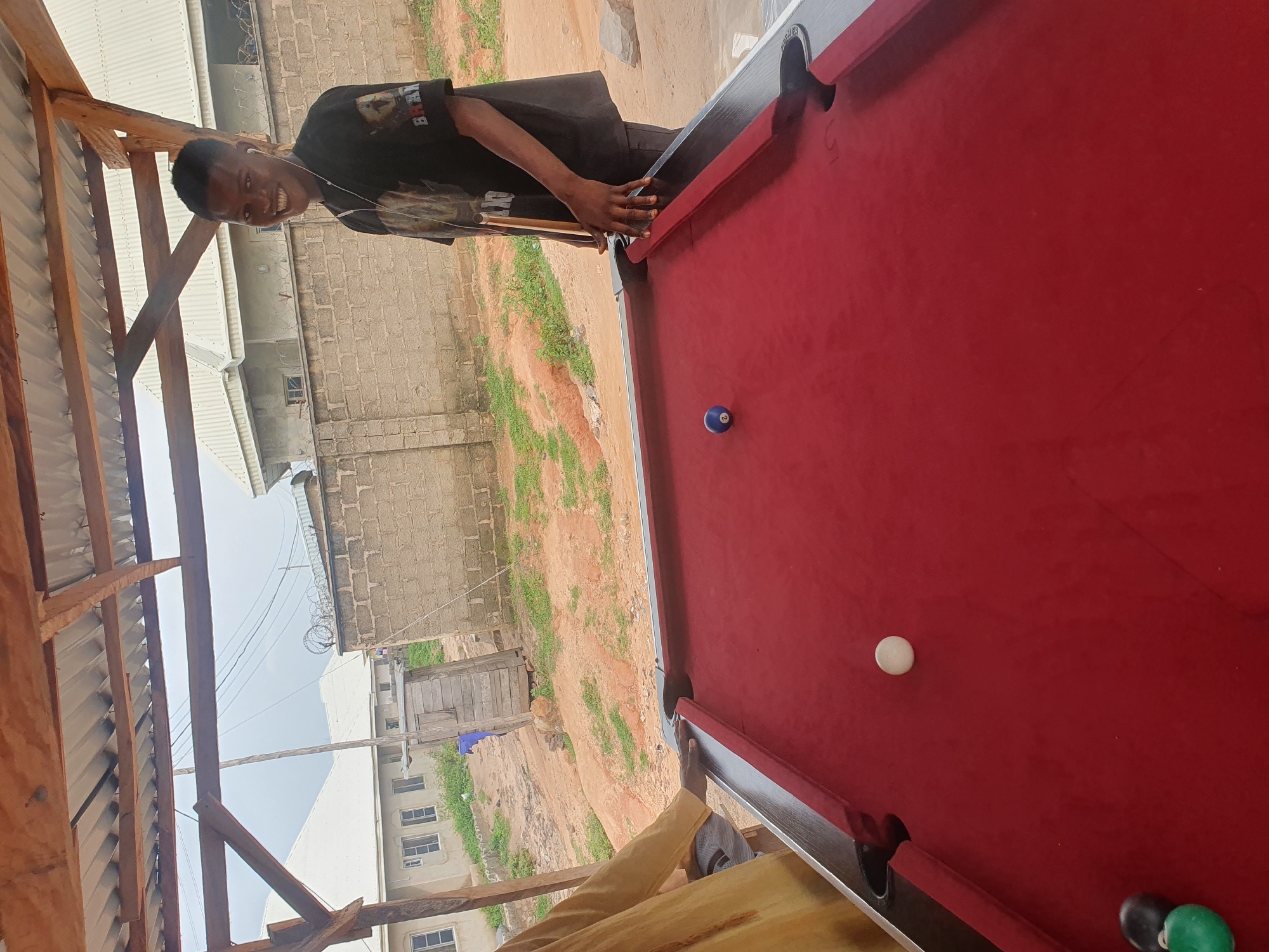 A young man in a black t-shirt and dark pants is standing on the right side of a red pool table, holding a pool cue in his right hand. He is looking towards the camera and smiling. He has white earbuds in his ears. The pool table has several balls on it: a white cue ball, a blue ball with the number 2, a white ball, a black ball, and a green ball. The table is outdoors, under a wooden and corrugated metal roof. In the background, there is a brick wall, a wooden structure, some buildings, and vegetation. The scene appears to be in a dusty, outdoor setting, possibly in Lokoja, Nigeria, given the style of architecture and the environment.