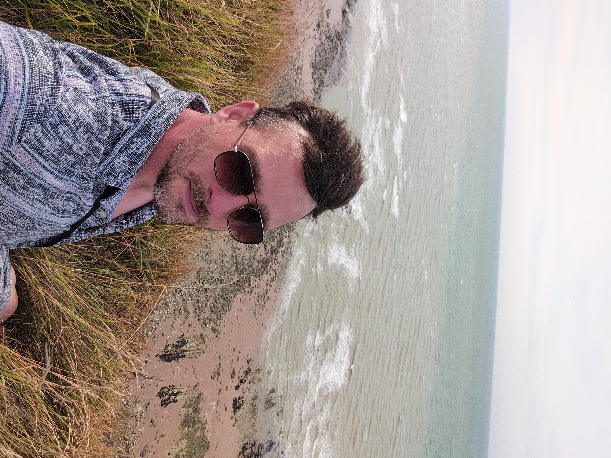 A man is taking a selfie on a grassy cliff overlooking the sea in Saint-Valery-en-Caux, France. He is wearing sunglasses and a patterned shirt. The sea is calm with small waves. The beach below is rocky and sandy. The sky is overcast.