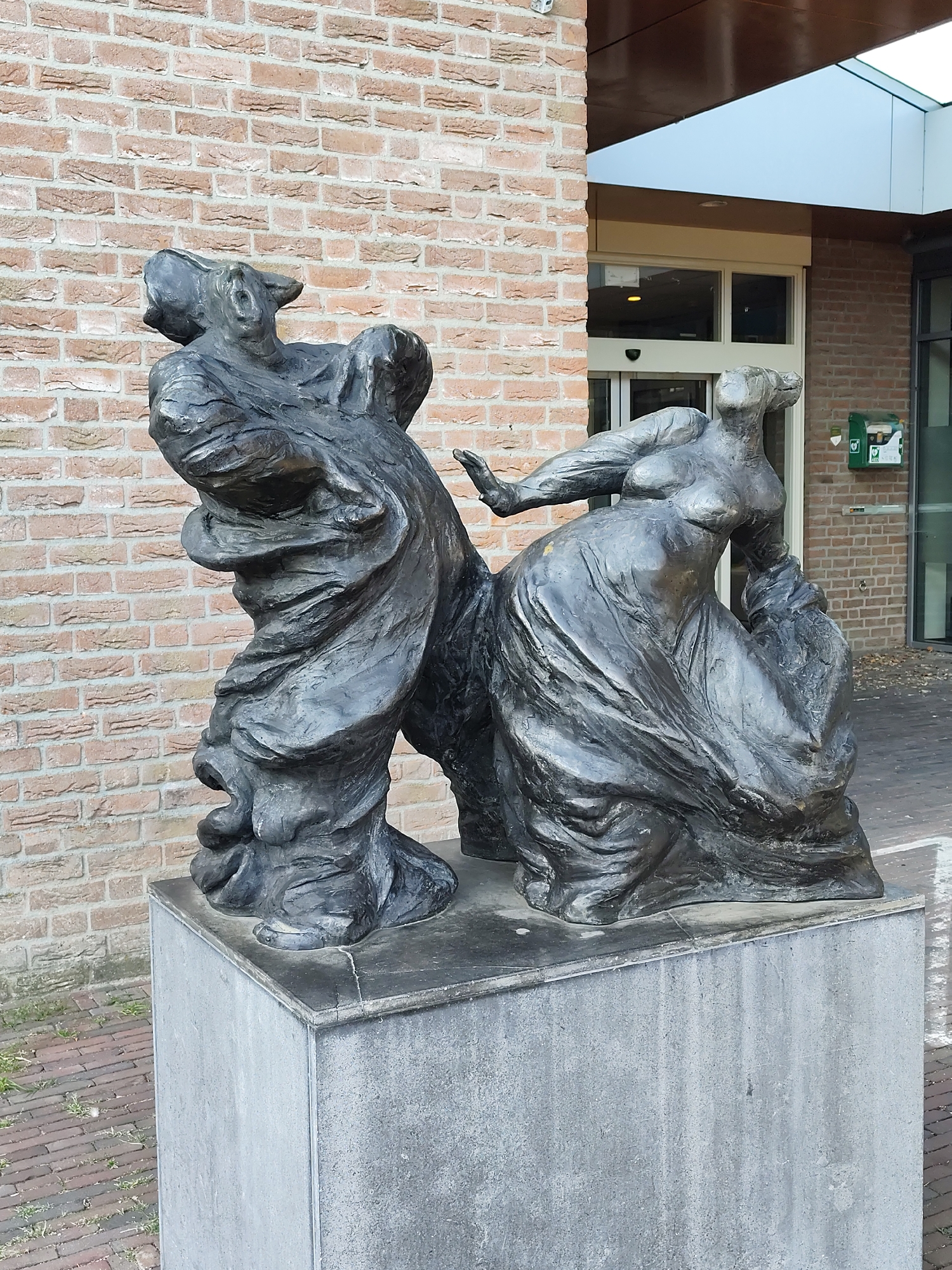 A bronze sculpture of two figures, likely depicting a dramatic scene, stands on a grey concrete pedestal. The figures are draped in flowing garments that obscure much of their bodies, but appear to be in dynamic poses, with one figure reaching out to the other. The sculpture is positioned in front of a red brick building with a modern entrance and a light-colored overhang. A green emergency medical box is mounted on the brick wall to the right of the entrance. The ground in front of the sculpture is paved with bricks. This sculpture is located in Schaijk, Netherlands.