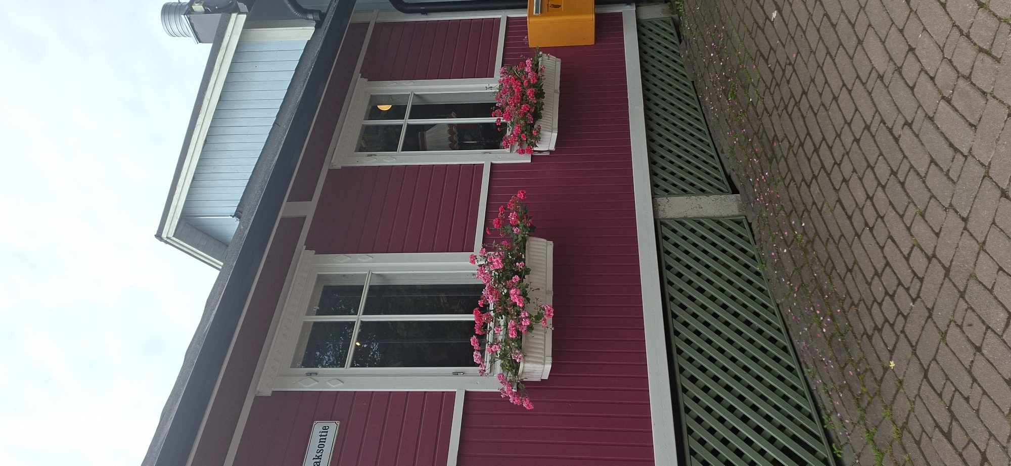 A close-up, eye-level shot captures the facade of a pink-painted wooden building in Naantali, Finland. Two white-framed windows are visible, each adorned with a white planter overflowing with vibrant pink flowers. The lower portion of the building features a green lattice structure, presumably a ventilation grate. To the right, a bright yellow mailbox is mounted on the wall. The ground in front of the building is paved with reddish-brown bricks laid in a herringbone pattern, with scattered pink flower petals and a few blades of grass. In the background, a light blue structure with white trim is partially visible, suggesting a neighboring building or an extension. The sky is overcast and pale. A street sign reading "Maksontie" is affixed to the left side of the building.
