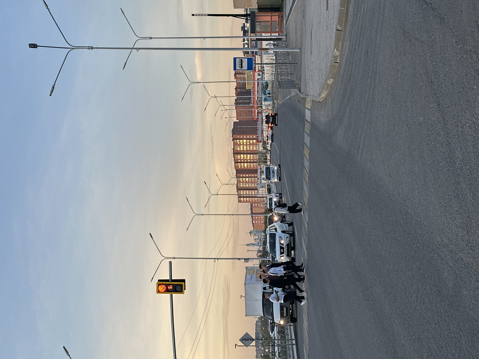 A street scene in Kokshetau, Kazakhstan, at dusk. A group of people are walking across a crosswalk. There are cars and trucks stopped in the street. A traffic light shows a countdown of 19 seconds remaining. Buildings line the street. There is a bus stop. Street lights are visible. The sky is light blue.
