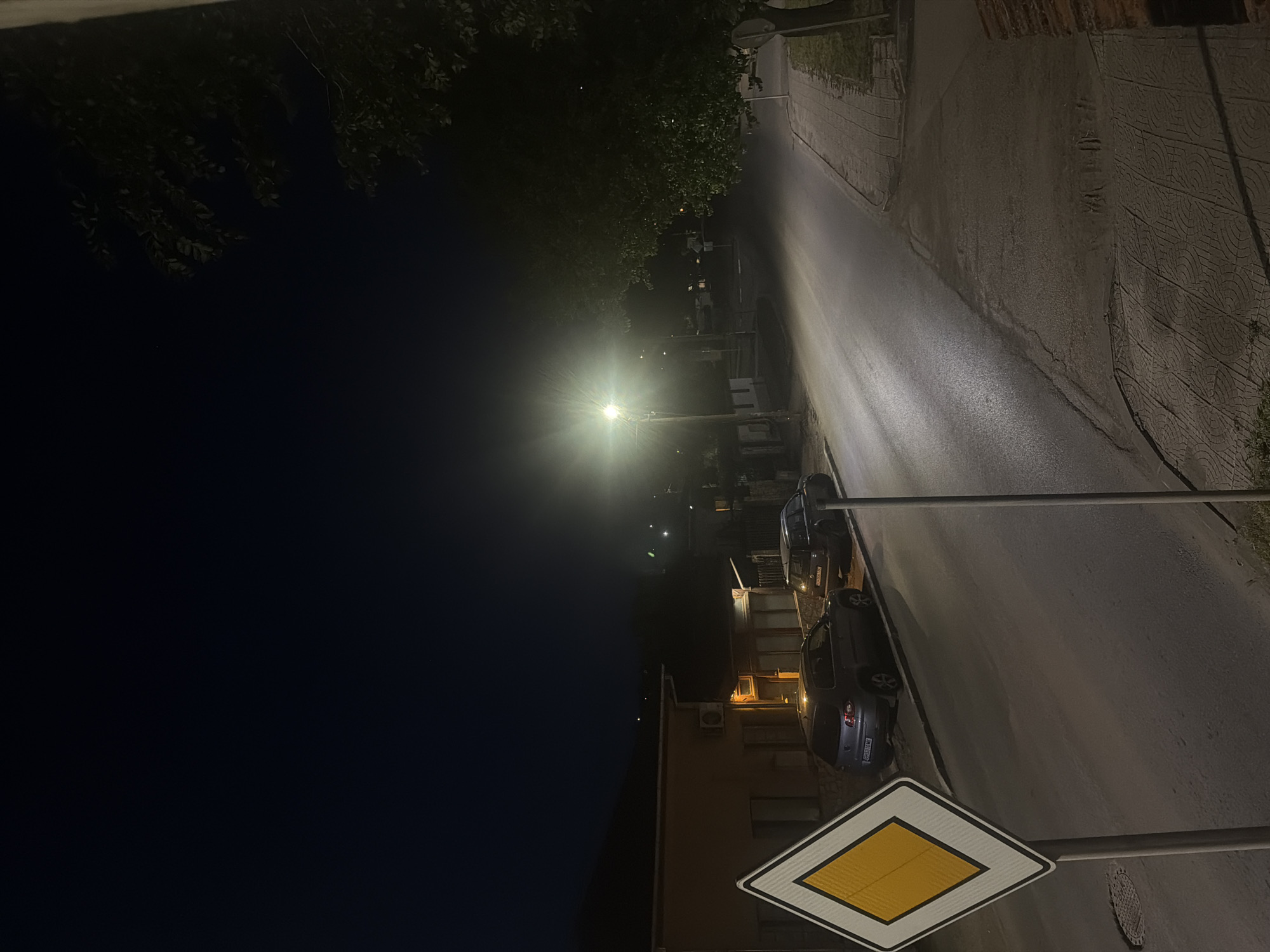 The image displays a quiet residential street at night, illuminated by a prominent streetlamp and ambient light from nearby buildings. The perspective is slightly elevated, looking down a gently curving asphalt road.
To the left, the dark night sky is partially obscured by the silhouettes of tree branches with visible foliage. A bright streetlamp acts as the central light source, emitting a strong glow that spreads across the road surface.
Along the right side of the street, a paved area or sidewalk is visible, bordered by a wall with decorative patterns. Two cars are parked along this side; the rear license plate of the closer vehicle reads "CB 4531 MW." Behind the parked cars, a two-story building with illuminated windows and an air conditioning unit suggests residential occupancy.
In the immediate foreground, a white square road sign with a yellow diamond, indicating a priority road, is clearly visible. The sign is mounted on a metal pole. Further down the street, beyond the primary light source, other smaller light points are visible in the distance. The overall scene conveys a peaceful, calm atmosphere typical of a residential area after dark. The time of day is clearly night, with no natural light visible.
