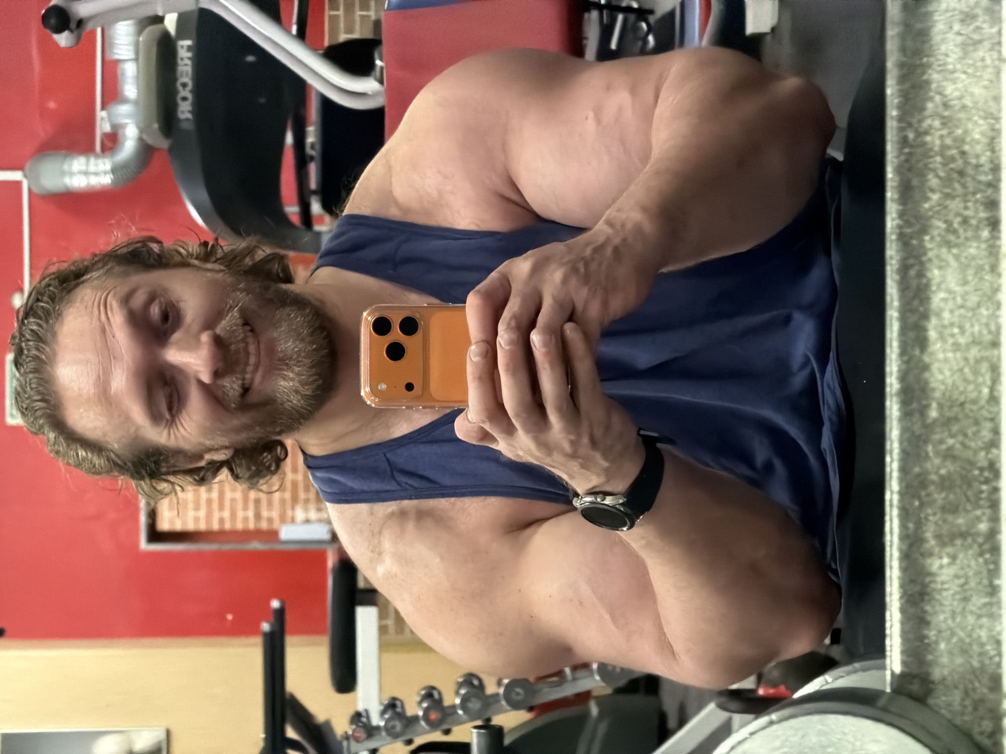A man is taking a selfie in what appears to be a gym. He is wearing a blue tank top and a watch on his left wrist. The gym contains exercise equipment, including weights and machines. A red wall is visible in the background. The location in the city of Saint Petersburg, Russia, cannot be determined based on the visual information provided.