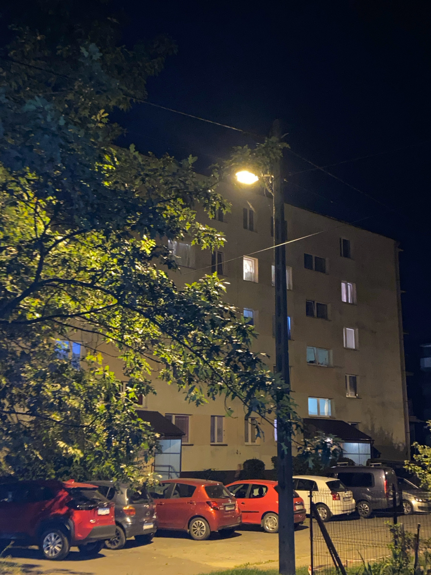 The image depicts an apartment building at night, likely in the city of Nowy Sącz, Poland. A street lamp illuminates a portion of the building and the surrounding area. Several cars are parked in front of the building. A large tree with green leaves is prominently featured in the foreground, partially obscuring the view of the building and the cars. The windows of the apartment building show some lights, indicating that the building is occupied. The sky is dark, suggesting it is nighttime. The scene appears to be an urban residential area.