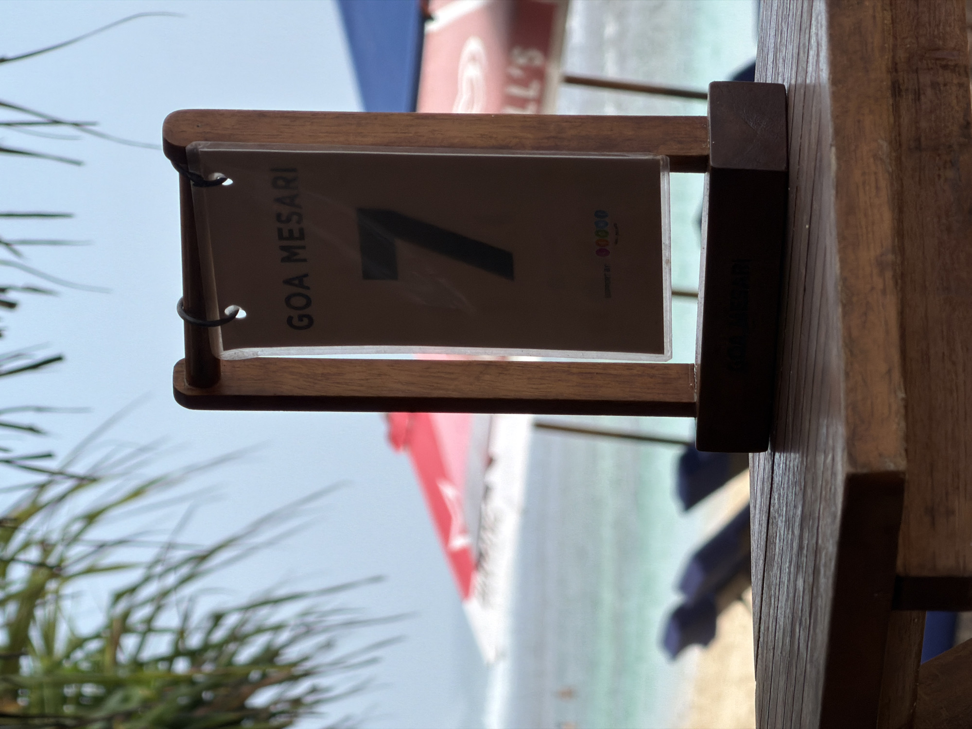 The image shows a table number sign on a wooden table in front of a beach in Kuta Selatan, Indonesia. The sign is made of wood with a clear plastic cover displaying the words "GOA MESARI" and the number "7". In the background, there are two beach umbrellas, one red and white, and the other blue. There are also beach chairs and the ocean. Palm fronds are visible in the upper left corner of the image.