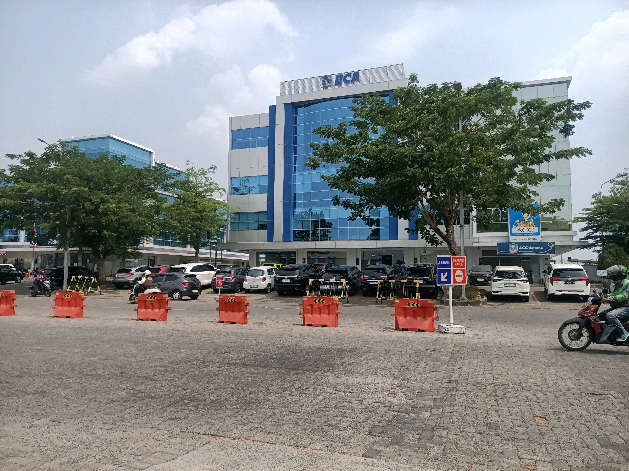 In Grogol, Indonesia, a paved exterior lot features several orange traffic barriers lined up in the foreground. Beyond these barriers, a row of parked cars, mostly dark-colored sedans and SUVs, are visible in front of modern buildings. One building, with a blue and white facade and large glass windows, displays the "BCA" logo at the top. Another building to the right has signage that includes "ACC" and "member of ASTRA". A person on a red motorcycle, wearing a green jacket and helmet, rides across the lot towards the right side of the frame. Trees are strategically planted around the buildings and lot, and the sky is partly cloudy.