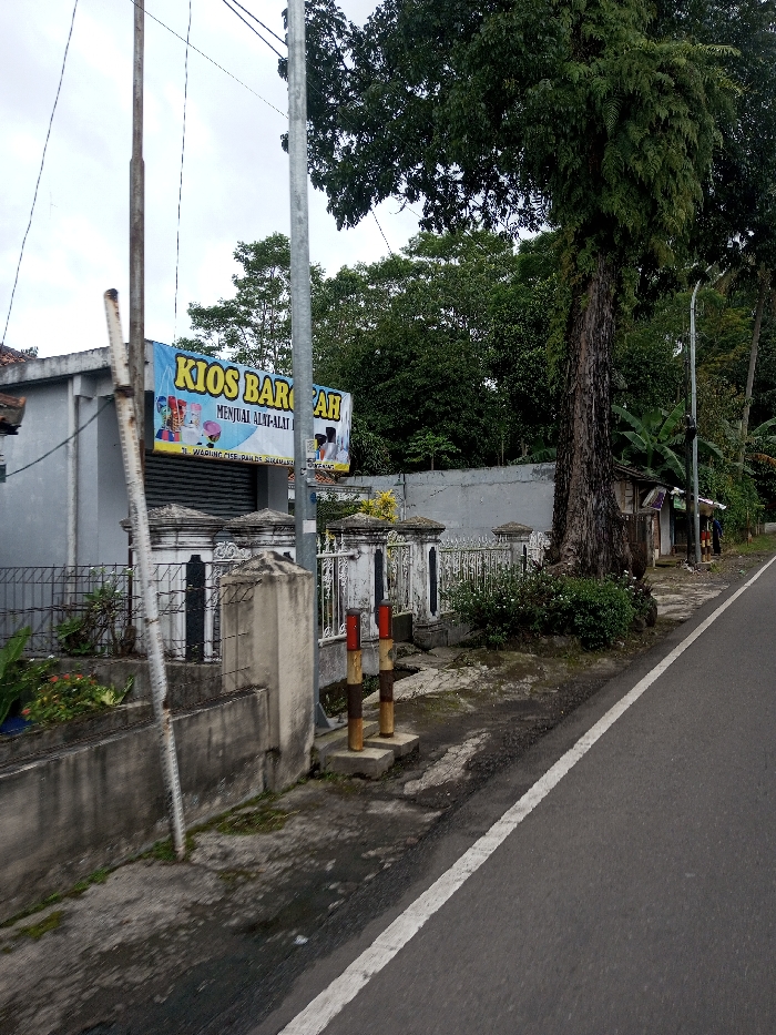 The image depicts a roadside scene, identified by a banner as "JL. WARUNG CISE, KAB. BANDUNG," placing the location in Bandung, Indonesia. The sky is overcast.
In the left foreground, a low concrete retaining wall with sparse vegetation leads to a decorative, white and black concrete and metal fence. Behind this fence is a light-grey, single-story building with a closed corrugated metal roll-down door. A blue banner prominently displayed on the building reads "KIOS BAROKAH" in yellow letters, with "MENJUAL ALAT-ALAT..." (Sells tools/equipment...) and the aforementioned address below.
Multiple utility poles are present; a rusty metal pole stands on the far left, and a grey metal pole is centered in the midground, both supporting overhead lines. To the right of the building and fence, a large tree with a textured trunk and dense green foliage dominates the midground. Behind the fence and tree line, a grey concrete wall is visible, backed by lush green trees and vegetation.
The foreground features a grey asphalt road with a solid white line marking its edge. A dirt and muddy strip runs parallel to the road, containing two short, concrete-based bollards painted with alternating red/white and brown/yellow bands. Further down the road on the right, additional informal structures are visible, and two indistinct human figures are present in the distance. A street light pole is also visible further down the road on the right.