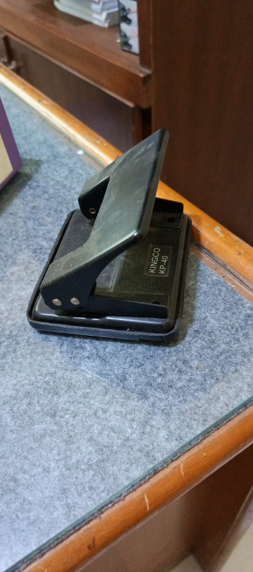 The image shows a black hole puncher on a grey textured surface. The hole puncher has the brand name "KINGCO" and the model number "KP-40" printed on it. The background appears to be a wooden surface with some books or papers on a shelf above. The scene is indoors. The location of Banda Aceh, Indonesia is not visually discernible from the provided image.