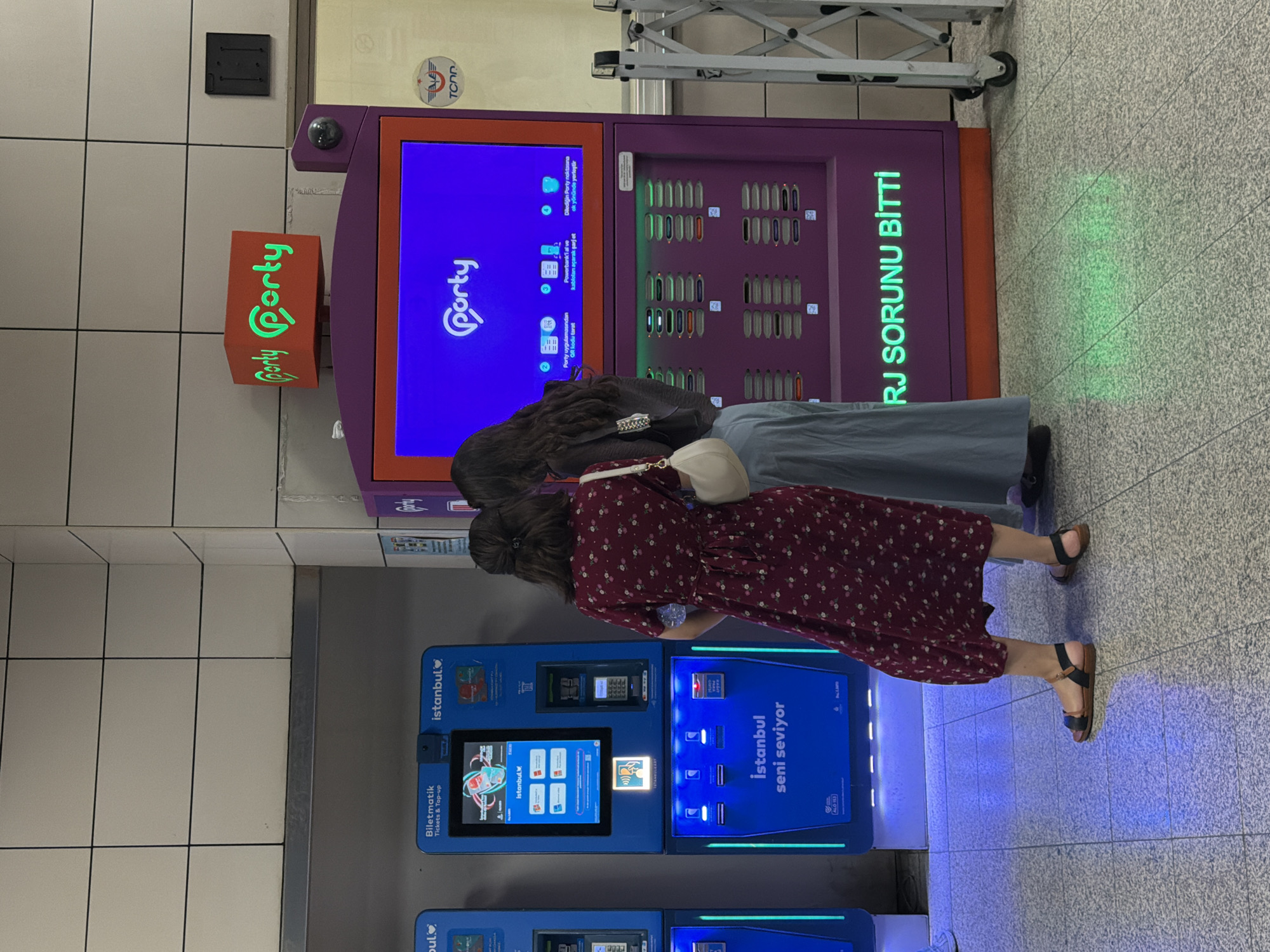 In Istanbul, Turkey, two women stand near a purple charging station and two ticket machines at a subway station. The woman on the left wears a dark red floral dress and black sandals. The woman on the right wears a grey top and a light blue skirt. The charging station displays a blue screen with the "Porty" logo and charging instructions in Turkish. The ticket machines are blue and feature the "Istanbulkart" logo. The floor is tiled and illuminated with blue light from the machines. A silver gate is visible to the right of the charging station.