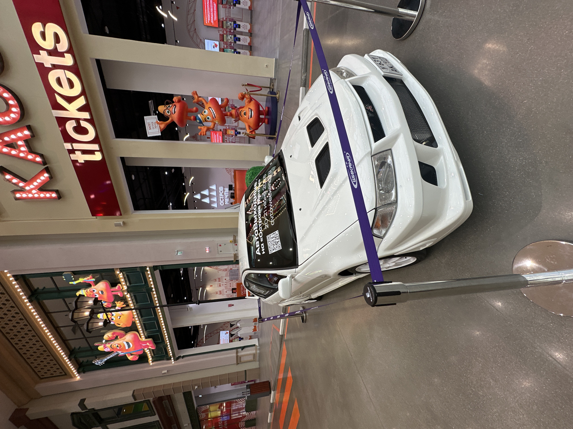 A white, customized sports car is displayed indoors within a large entertainment complex or shopping mall, identified as "Dream Island" in Moscow, Russia. The car, possibly a Mitsubishi Lancer Evolution given its styling, is cordoned off by purple stanchion ropes, which are marked with "ШИКАНА" (Shikana). A sign on its windshield advertises an "АвтоВыходные на «Острове Мечта»" (Auto-Weekend at Dream Island) event scheduled for June 20-21, 2026, accompanied by a QR code. The partially visible license plate reads "АК 97" and "Y631".
In the background, a prominent illuminated sign above an entrance reads "tickets" in English, with partially visible Russian text "КЛЮЧЕ НАЧИНАЮ" (Klyuche Nachinayu). Several reddish-orange, peanut-shaped cartoon characters adorn the space; two are near the car, one holding a "БИЛЕТ ОСТРОВ МЕЧТЫ" (Ticket Dream Island) sign, while three others hang from the ceiling, depicted playing musical instruments. Further "ОСТРОВ МЕЧТЫ" (Dream Island) signage is also visible. The event takes place in a brightly lit indoor environment with a grey floor featuring orange stripes.
