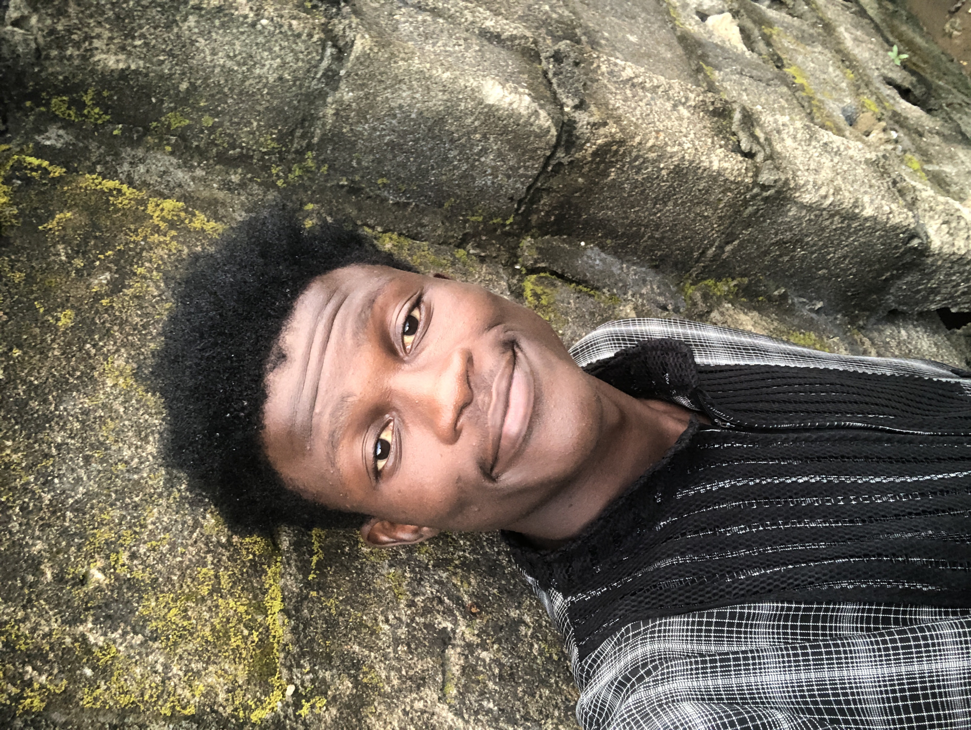 A young Black male is shown from the chest up, taking a selfie against a textured, moss-covered stone wall. He has short, dark, curly hair and is smiling at the camera. He is wearing a black and white plaid shirt with a black mesh panel on the chest and vertical striped detailing. The background is a rough, uneven stone wall with patches of green moss. The image appears to have been taken outdoors, possibly in a natural or urban environment with exposed stonework.