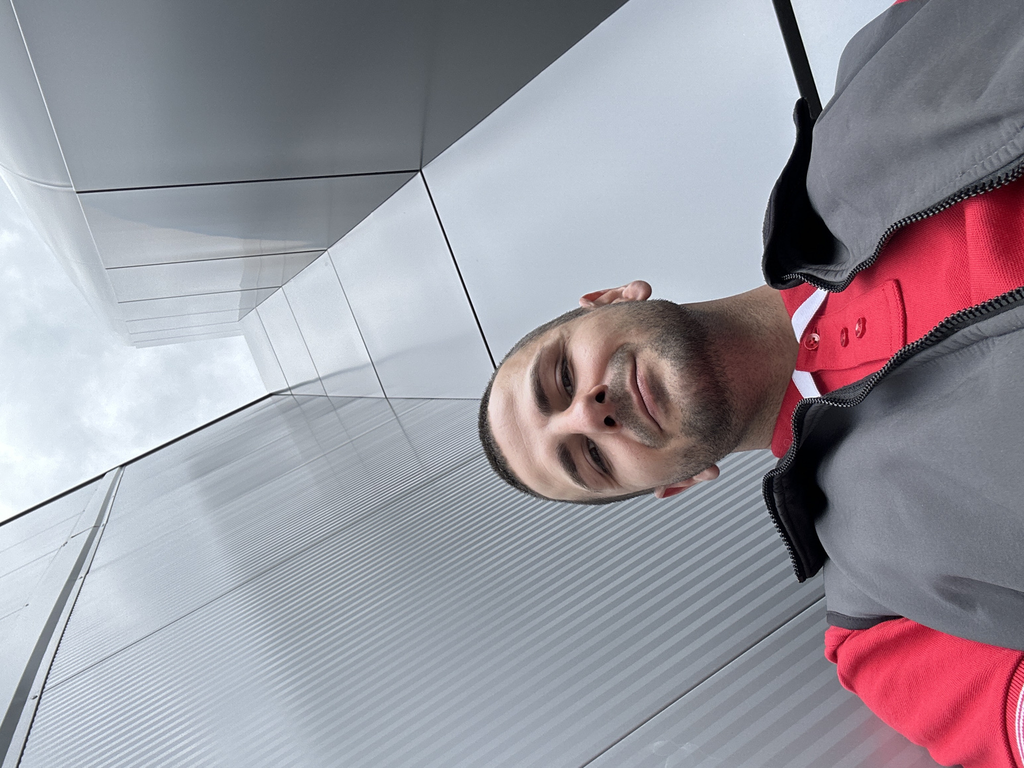 A low-angle selfie of a man with short brown hair and a trimmed beard, wearing a red collared shirt and a gray vest, is positioned in front of a building with a gray metal facade featuring horizontal striations. The building is located in Lovech, Bulgaria. The architecture also includes sections with smooth, flat gray panels. The sky is overcast.