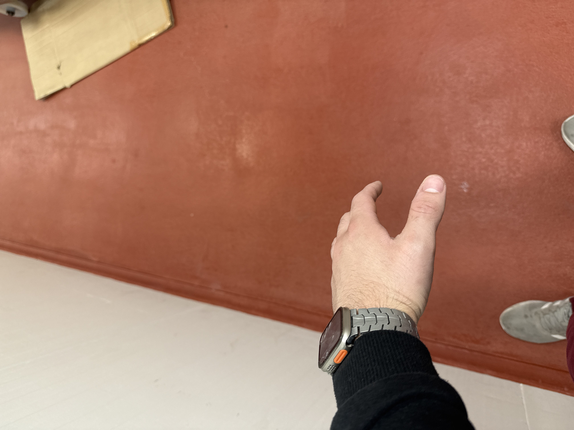 The image shows a first-person view of a person standing in a building, likely an interior space, in Stara Zagora, Bulgaria. The person is wearing a black sweater and a silver-colored smartwatch with a metal band. The floor is divided into two sections: one with white tiles and another with a reddish-brown painted surface. A corner of a yellow object is visible in the upper left. The person's hand is reaching out towards the reddish-brown surface. A portion of a shoe is visible in the lower right corner.