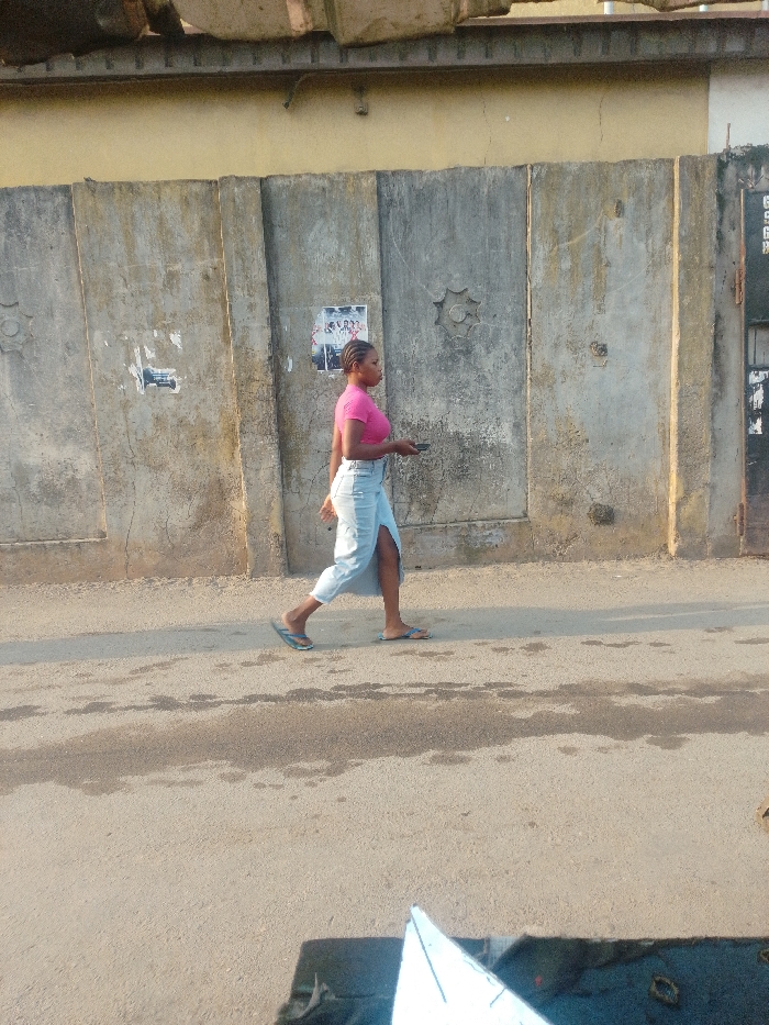 A young woman in a pink top and light blue denim skirt walks along a paved road. She's wearing sandals and holding a mobile phone. Behind her is a tall concrete wall with a faded yellow top. The wall has a few posters or signs on it and some decorative elements. The road surface appears worn, with patches and variations in texture. The photo appears to be taken during the daytime with natural lighting.