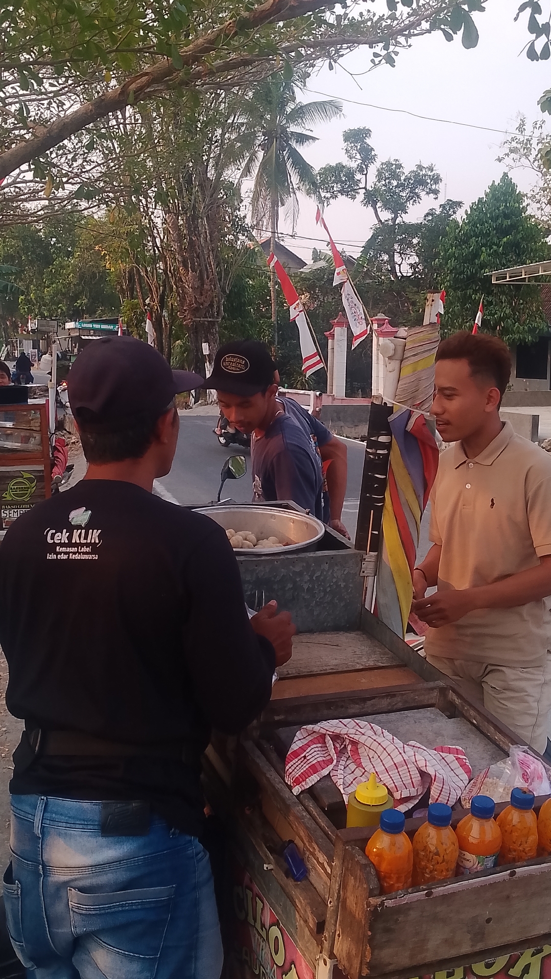 This image captures a street scene in Karanglangu, Indonesia, featuring a food cart and three individuals. The cart, constructed from wood, displays a variety of food items and condiments. In the foreground, a person wearing a black t-shirt with a white logo that reads "Cek KLIK" and blue jeans stands with their back to the camera. To their right, another person wearing a blue t-shirt and a black cap is leaning over the food cart, appearing to interact with the person in the black t-shirt. A third individual, wearing a tan polo shirt and light-colored shorts, stands to the right, engaged in conversation. The background reveals a street with passing traffic, including a motorcycle, and foliage, with Indonesian flags decorating poles along the roadside. The overall setting suggests a bustling outdoor commercial environment during daylight hours.
