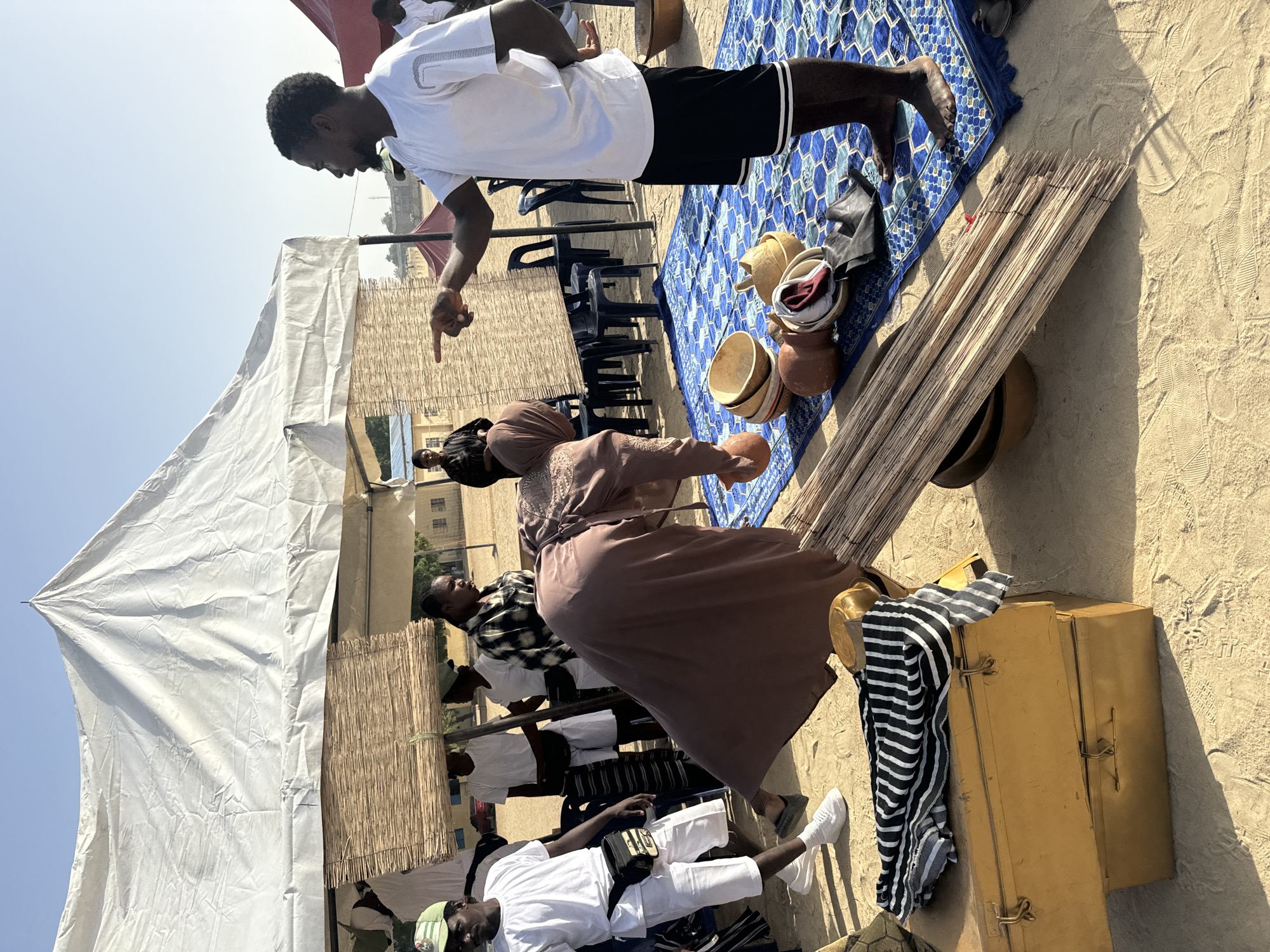 A bustling outdoor scene unfolds under a white tent structure, against a clear sky. A man in a white shirt and black shorts points downwards with his right hand, his left arm extended to maintain balance. He stands on a blue patterned rug, which is next to a stack of long, thin, light-colored wooden poles. Adjacent to the poles are assorted earthenware pots and bowls.
A woman in a brown dress bends over, appearing to handle or examine one of the pots. Several other individuals are situated under the tent, dressed in a variety of casual clothing. To the left is a man reclining wearing a white shirt, white pants, and white sneakers and sporting a green hat. A yellow suitcase with hardware is placed near him.
The environment seems to be sandy. The background includes buildings, partially obscured by the tent and the people in the foreground. Based on available information, it is likely that the location is in the city of Maiduguri, Nigeria.