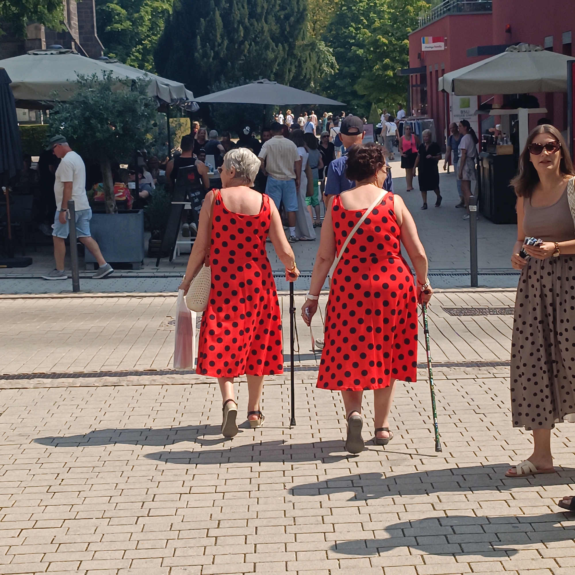 In the city of Bad Nauheim, Germany, on a bright, sunny day, two women are walking away from the camera on a paved pedestrian walkway. The woman on the left wears a red dress with black polka dots and carries a white mesh bag in her left hand. She uses a cane in her right hand. The woman on the right also wears a red dress with black polka dots and a white strap over her right shoulder. In the background, other pedestrians are visible, some sitting at outdoor cafes under umbrellas. To the right, another woman wearing a polka dot skirt and a neutral-colored top stands facing the camera, looking to her left. The environment suggests a commercial or recreational area with shops and eateries.