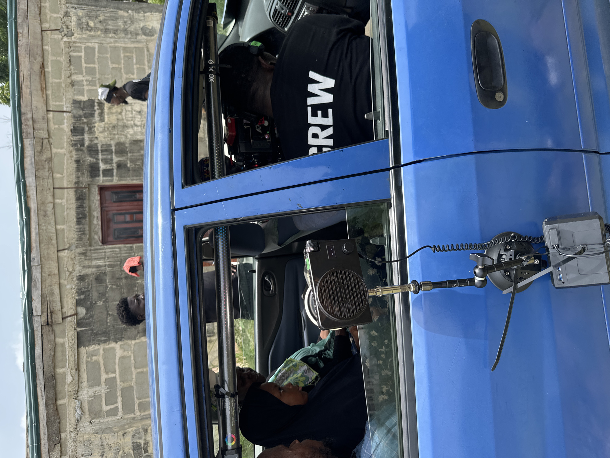 The image shows the interior of a blue vehicle, possibly a van, parked in what appears to be a residential area in Lagos, Nigeria. Inside the vehicle, there is a person wearing a black "CREW" t-shirt in the driver's seat, facing towards the front, and operating a camera. Another person is visible in the back seat. A rectangular device is attached to the outside of the van's window, possibly for recording sound. Outside the vehicle, people can be seen standing, partially visible. The background shows buildings constructed with gray brick.