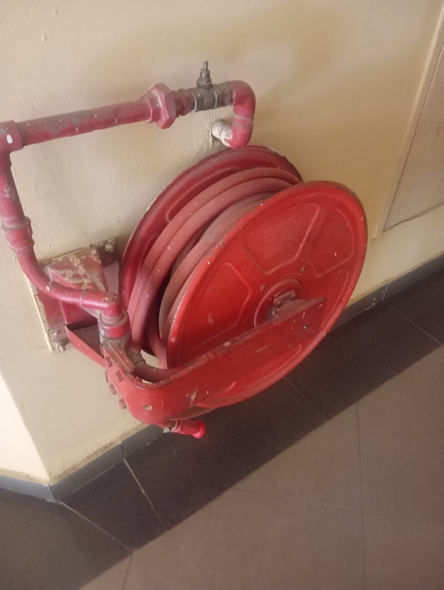 The image shows a red fire hose reel mounted on a pale yellow wall. The reel is made of metal and is coiled with a red hose. A red pipe connects the reel to a valve on the wall. The fire hose reel appears to be old and slightly worn, with some paint chipped off. The wall has some marks and smudges. The floor is tiled with dark gray and beige tiles. The scene is indoors, likely in a public building or facility. The fire hose reel is a standard piece of safety equipment. The location in Jos, Nigeria, is not discernible from the image alone.