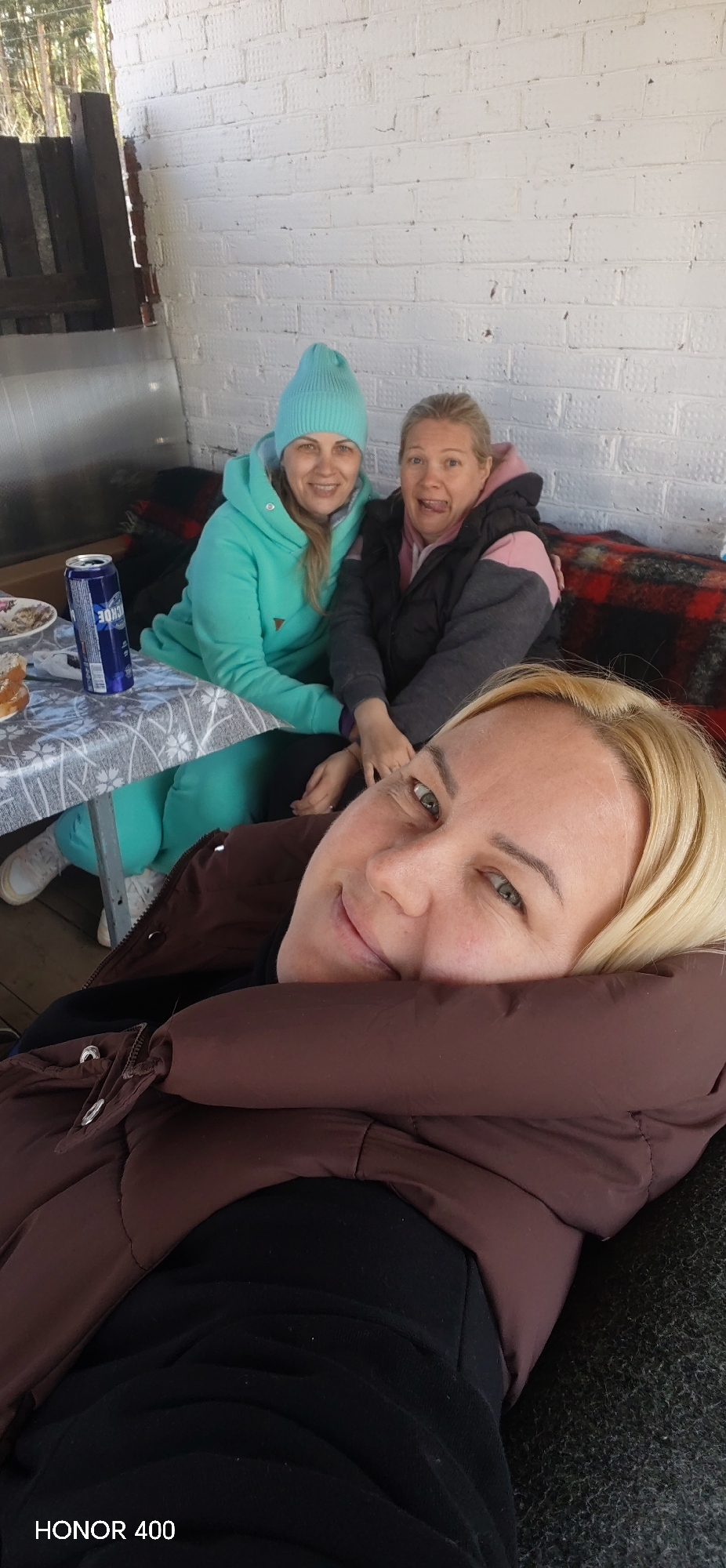 Three women are sitting on an outdoor patio. The woman in the foreground is blonde and wears a dark brown puffer jacket and black shirt. She is looking at the camera and smiling. Behind her, two other women are sitting on a bench covered with a red and black plaid blanket. The woman on the left wears a turquoise beanie and sweatsuit. The woman on the right wears a black puffer jacket with pink accents. There is a small table to the left with a can of beer and some food on it. The patio has a white brick wall in the background. A partially visible wooden structure is on the left. "HONOR 400" is written at the bottom left corner. The location is Noviy, Russia.