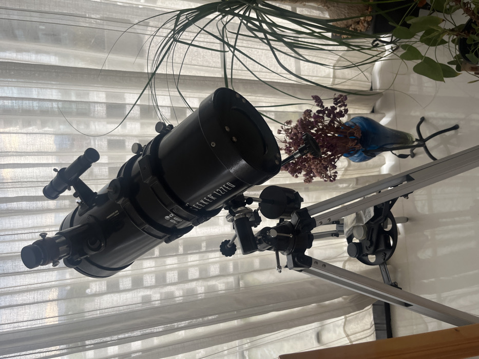 A black Celestron Seeker 127EQ telescope sits on a silver tripod. The telescope is pointed toward the left. Behind the telescope, a blue vase containing dried purple flowers is visible. A leafy green plant hangs above the vase. The background consists of a sheer white curtain.