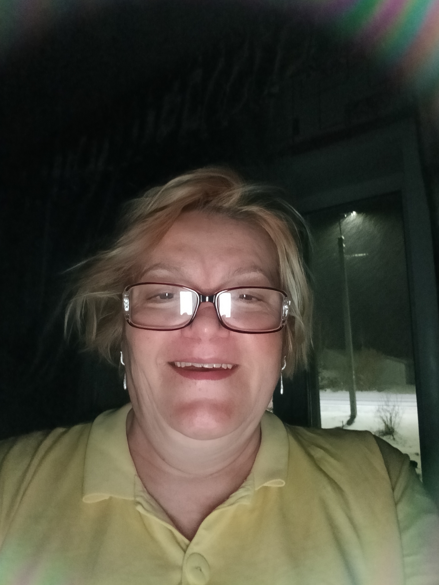 A person is centered in the frame, wearing glasses and a yellow collared shirt. The background is a mix of darkness and a visible area that appears to be outdoors at night, potentially during snowfall. A vertical pole or structure is visible, possibly a streetlight or signpost. The photo seems to be taken indoors, looking out. The location in the city of Красный Маяк, Russia, is not explicitly visible or confirmed within the image itself.